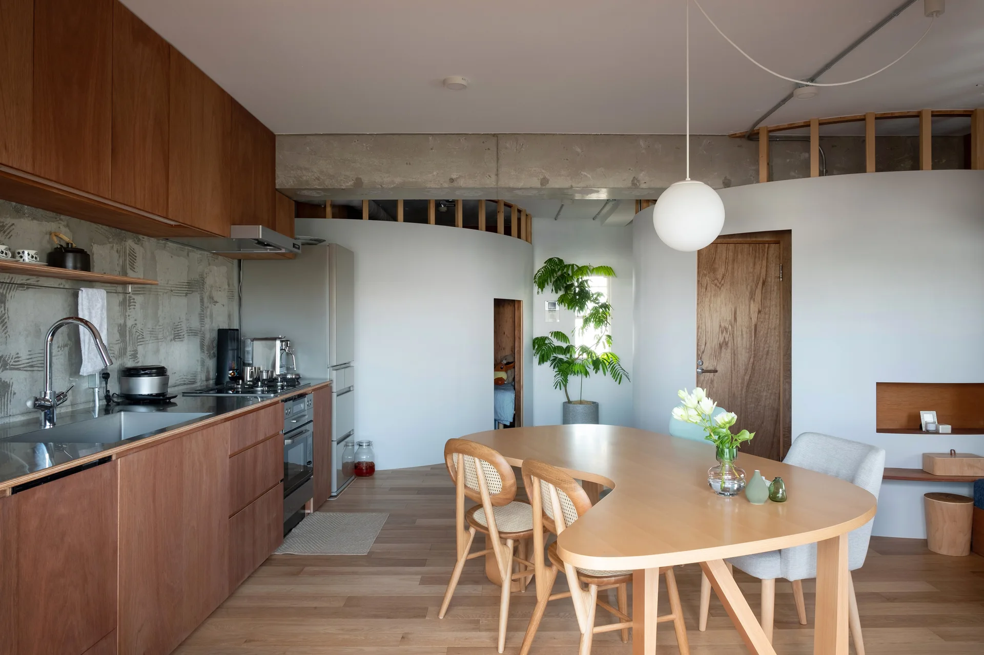 A modern open-plan kitchen and dining area features wood cabinets, a curved white partition wall, and a kidney-shaped wooden dining table.