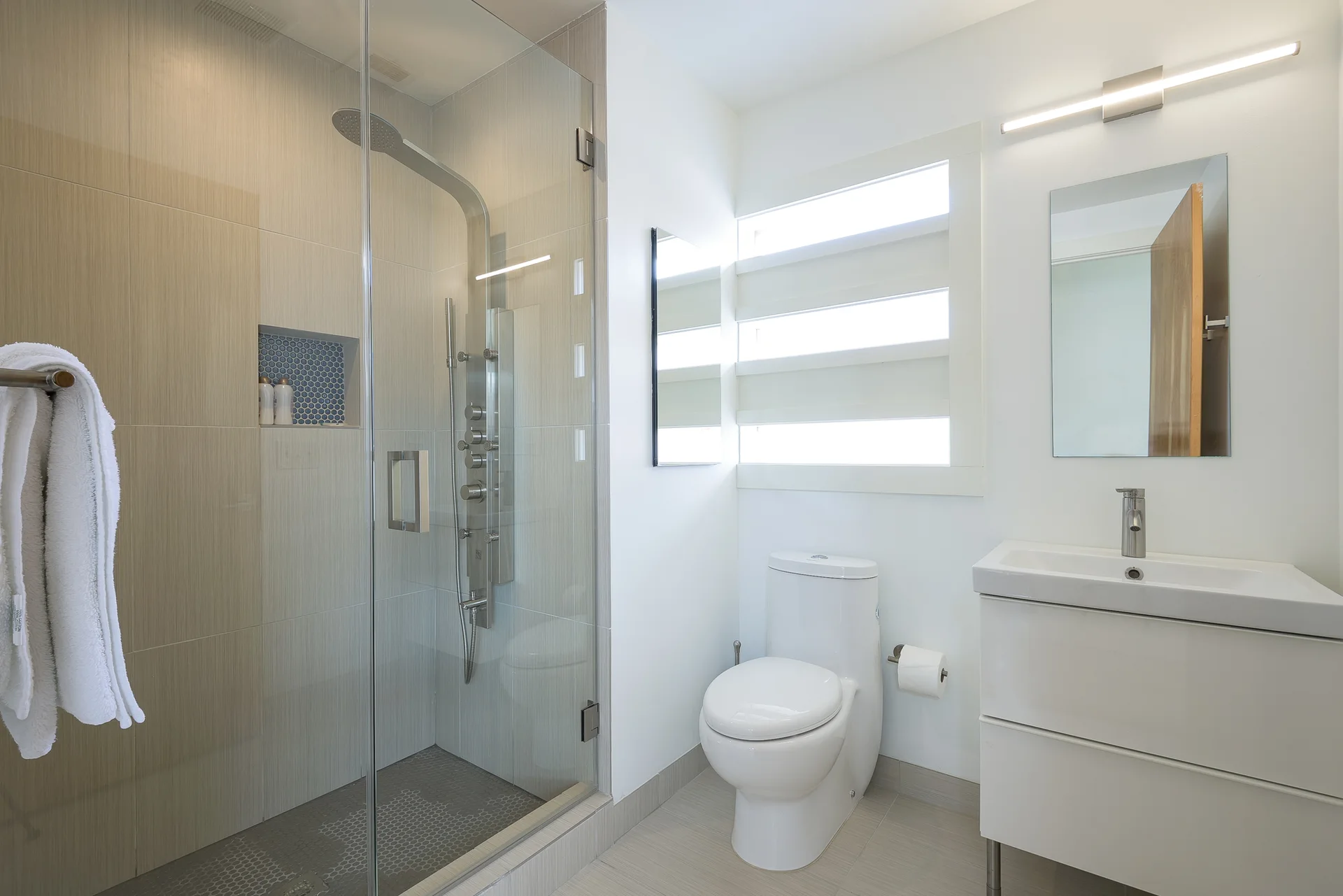 A modern bathroom featuring a glass-enclosed shower with beige tiling, a white toilet, and a sleek floating vanity with a rectangular mirror.