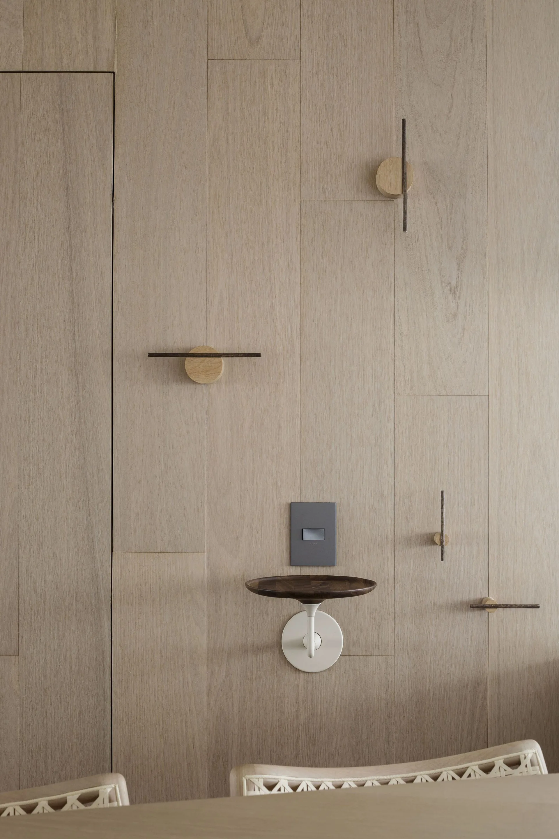 A light wood-paneled wall features minimalist geometric hooks, a grey switch plate, and a small wall-mounted circular shelf.