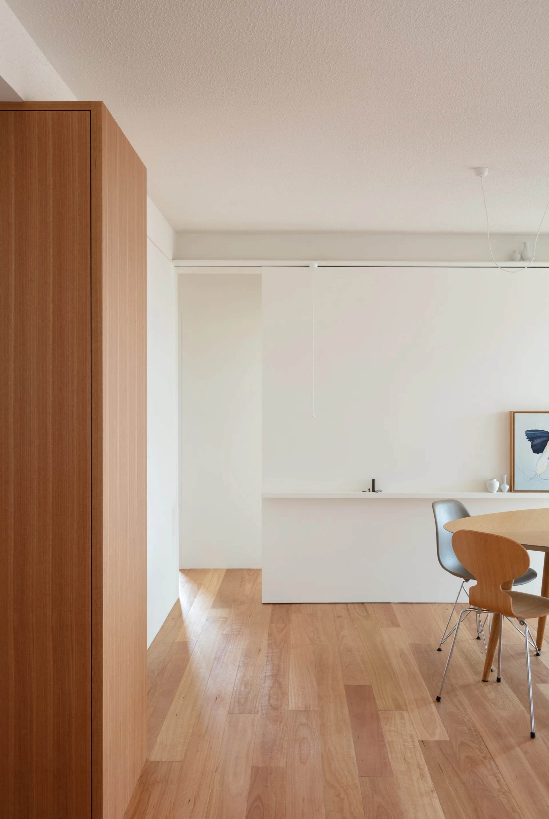 A minimalist dining room features a round wooden table, molded chairs, a low white partition wall with an integrated shelf, and light wood flooring.