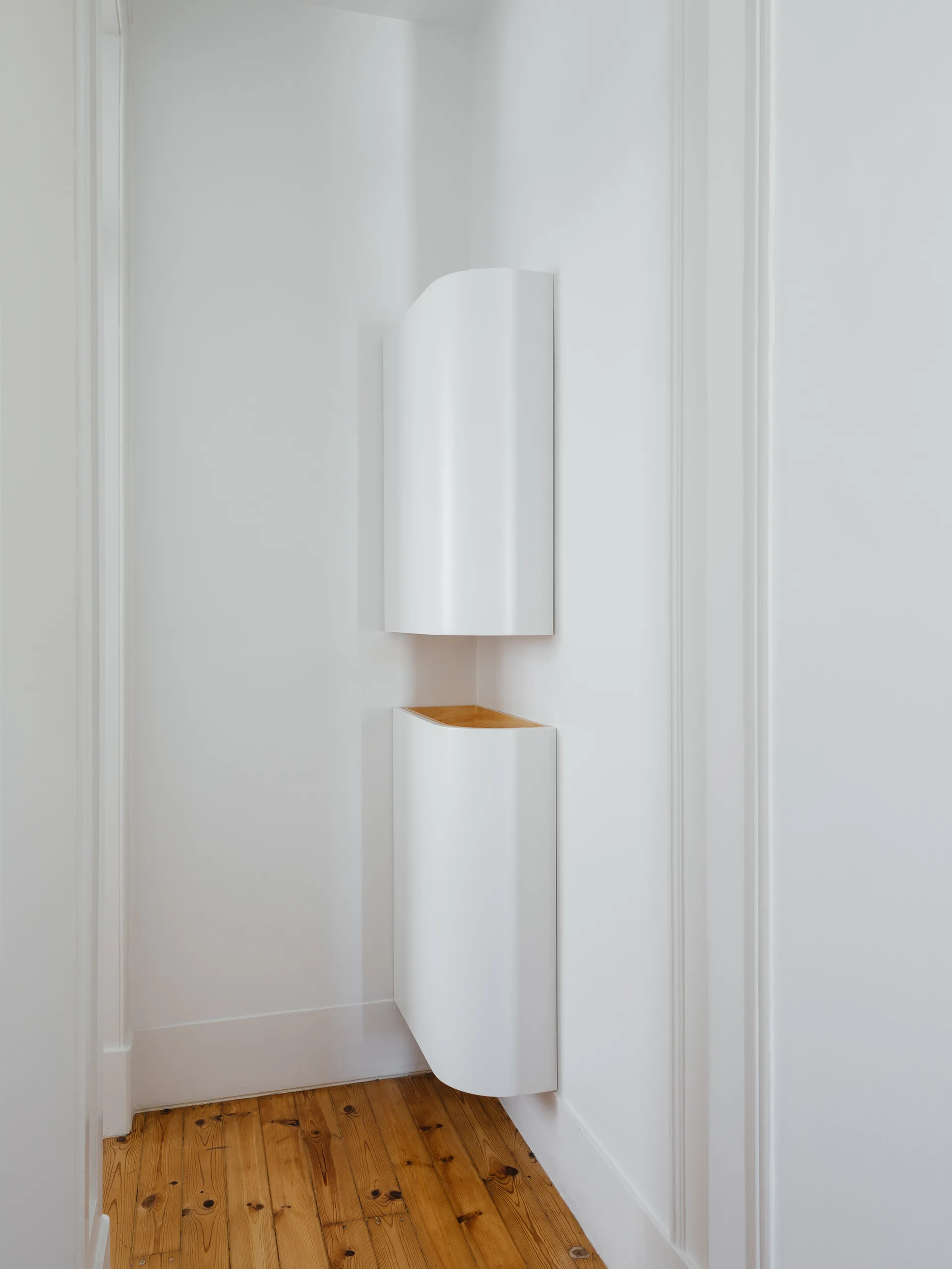Two stacked, white, curved corner cabinets are mounted on a plain white wall above natural pine hardwood flooring.