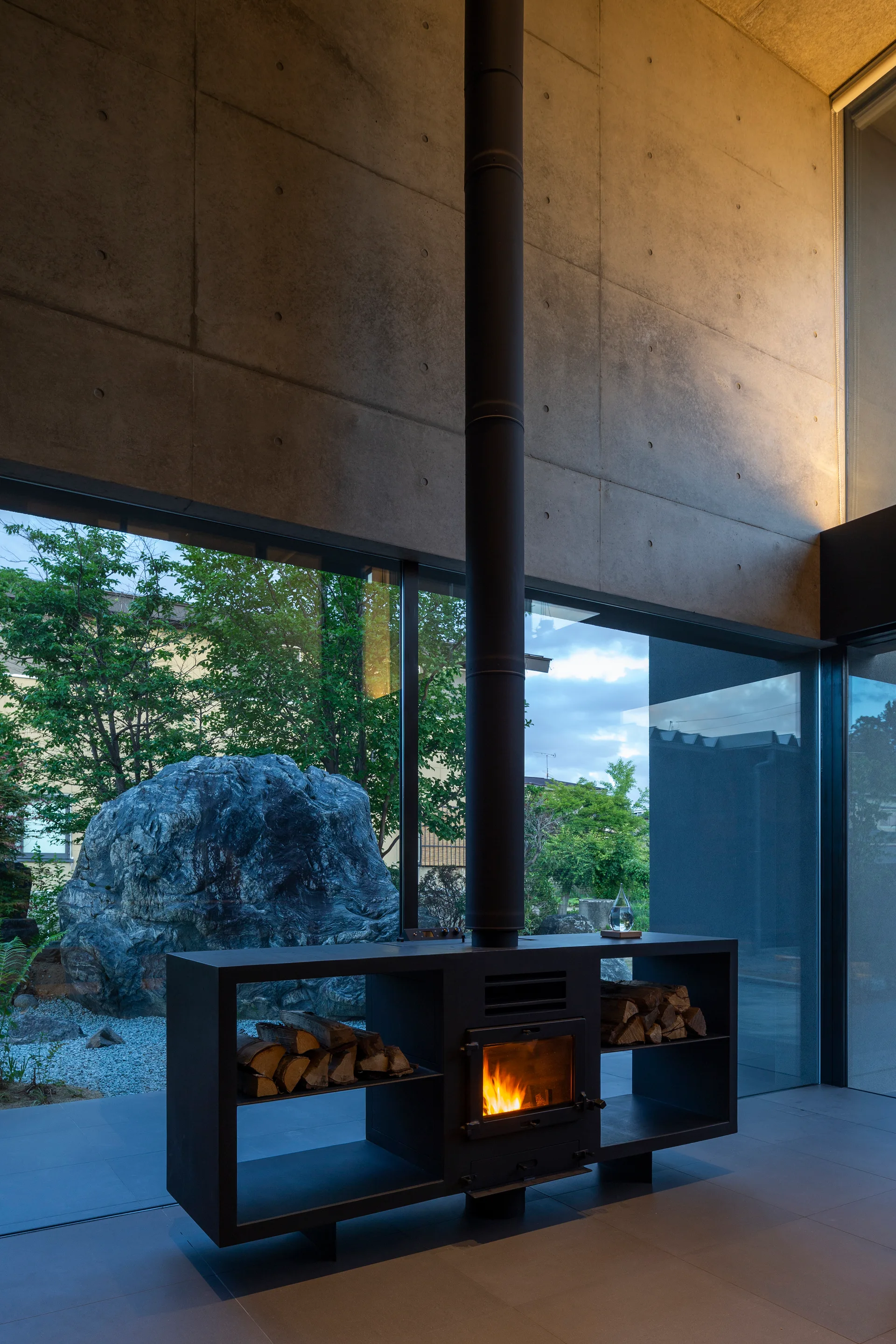 A black metal freestanding fireplace with built-in wood storage stands against an exposed concrete wall next to large floor-to-ceiling windows.