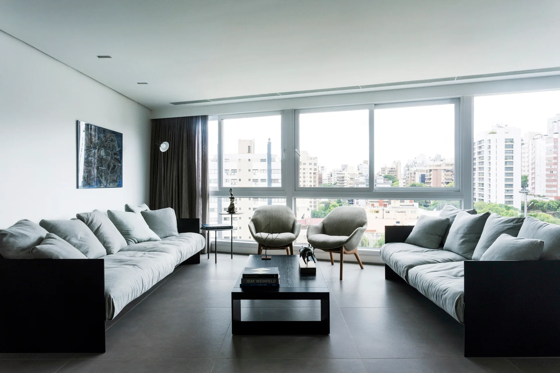 A minimalist living room features two light grey sofas facing each other, a black coffee table, and large windows showing a city.