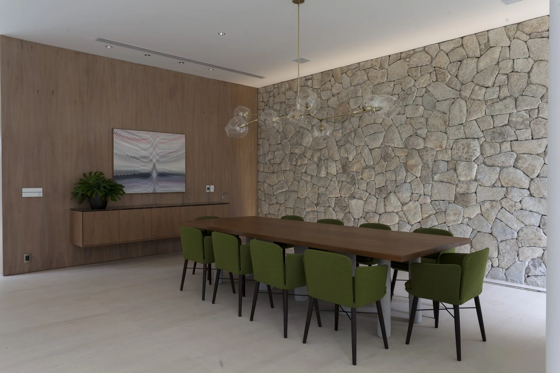 A modern dining room features a long wooden table, green upholstered chairs, a textured stone feature wall, and a branching glass chandelier.