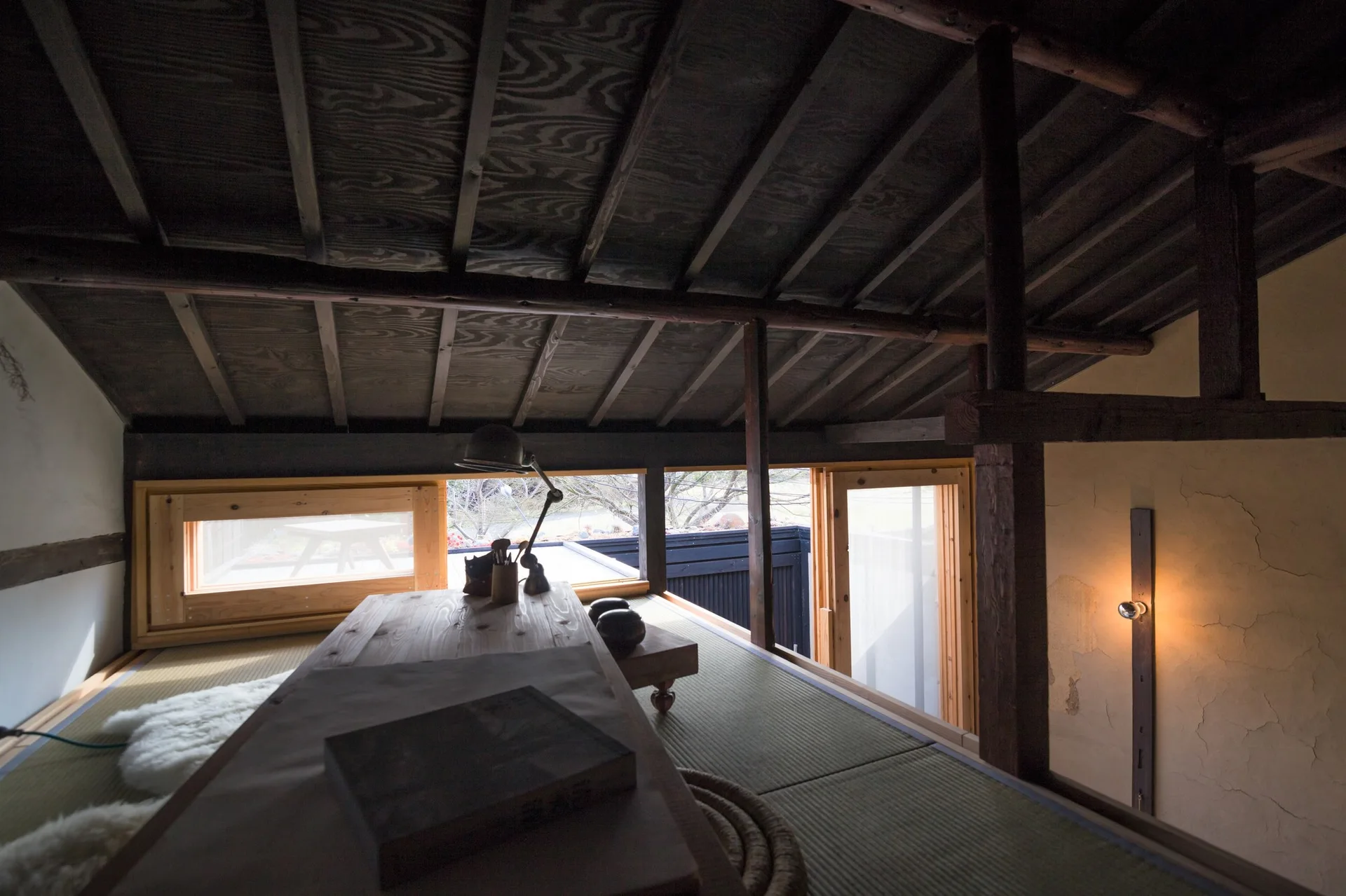 An attic loft features traditional tatami mat flooring under a sloped dark wood rafter ceiling with low horizontal windows.