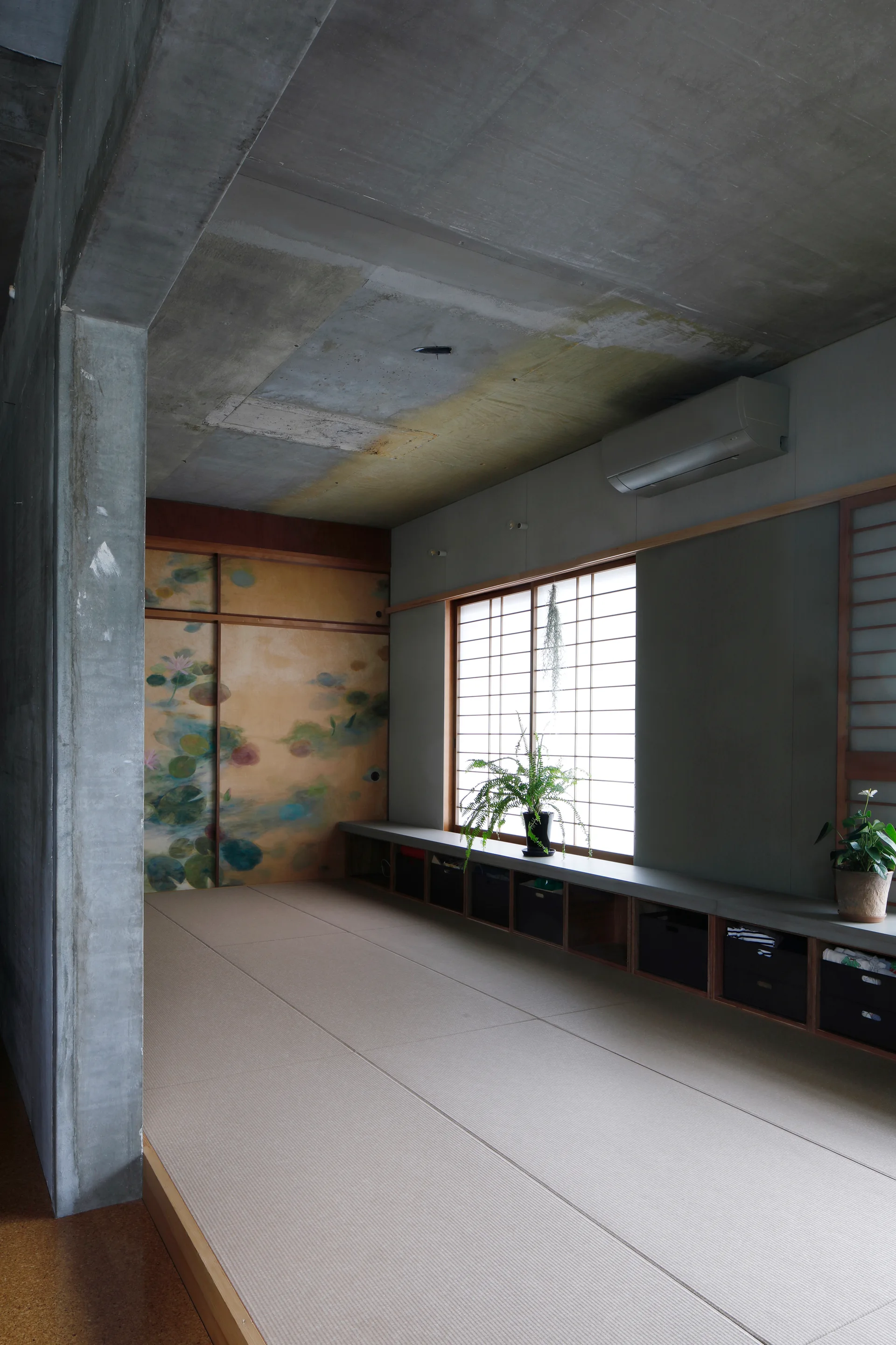 A minimalist room featuring tatami mat flooring, a raw concrete ceiling, shoji window screens, and a long low wooden storage bench.