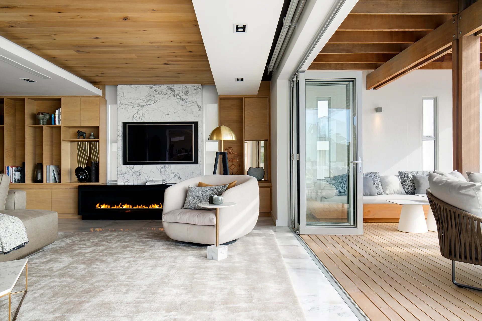A contemporary living room with a marble fireplace and wood ceiling opens through bifold glass doors to an outdoor wooden deck.