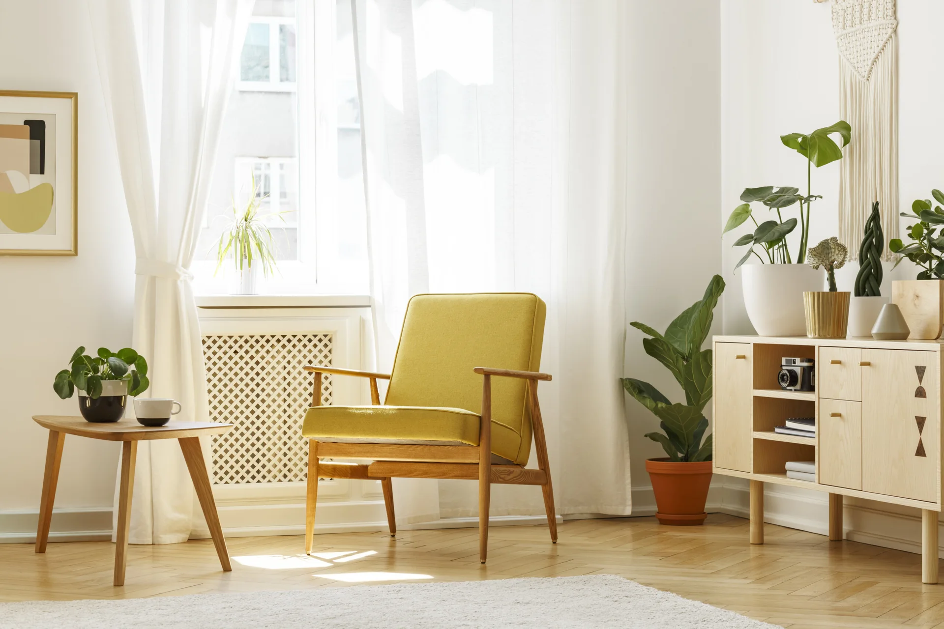A bright living room corner features a mustard yellow armchair, a wooden sideboard, and leafy houseplants on herringbone wood floors.
