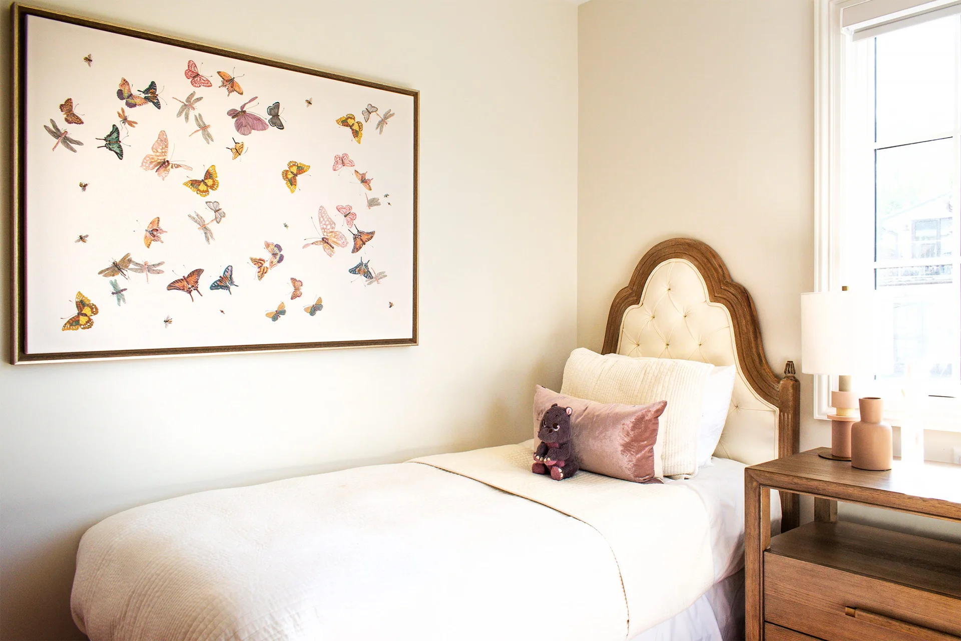 A twin bed with an arched tufted headboard sits beside a wood nightstand, below a large framed butterfly print on a cream wall.
