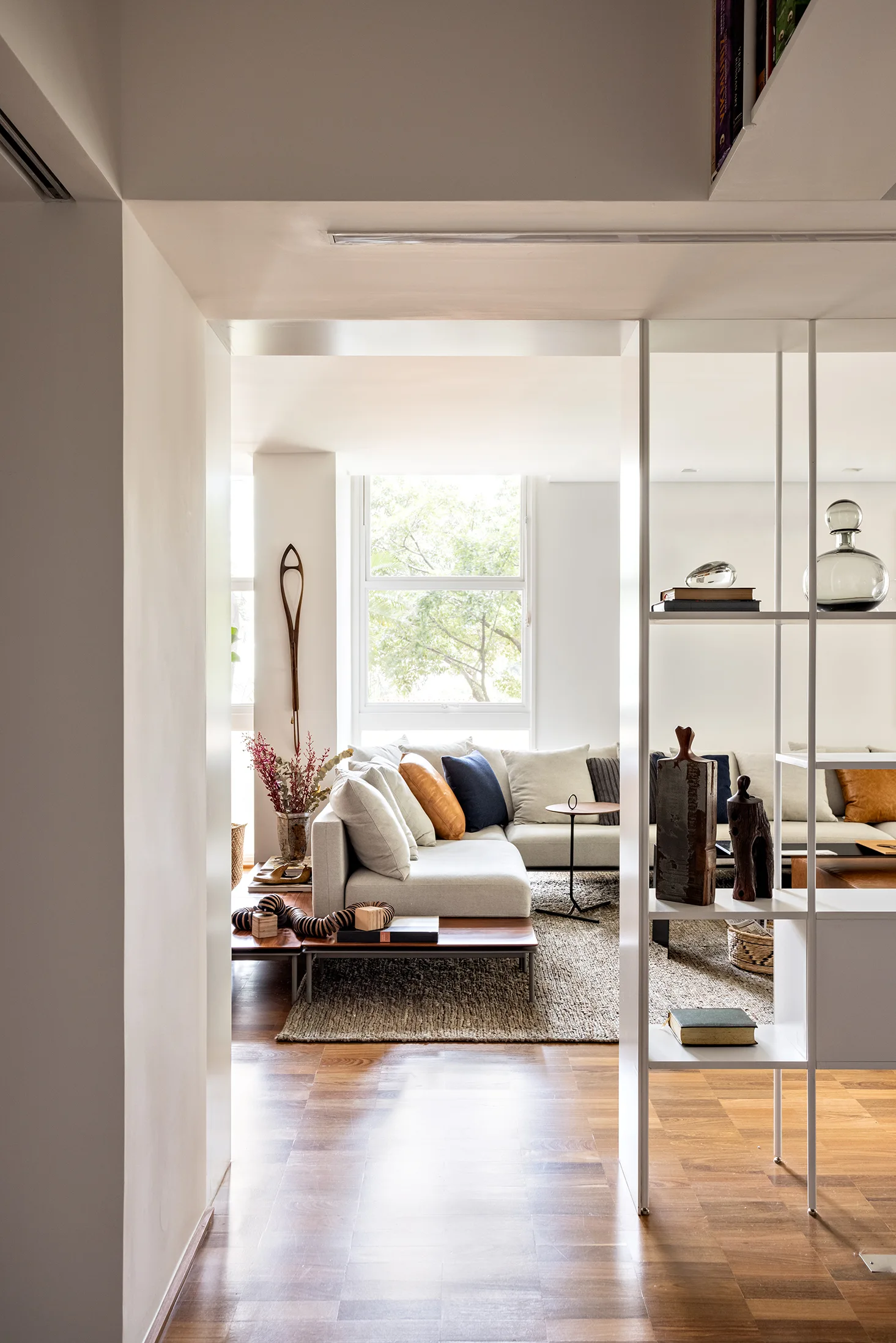 A light-filled living room featuring a low grey sectional sofa, a textured woven rug, and a white open shelving unit dividing the space.