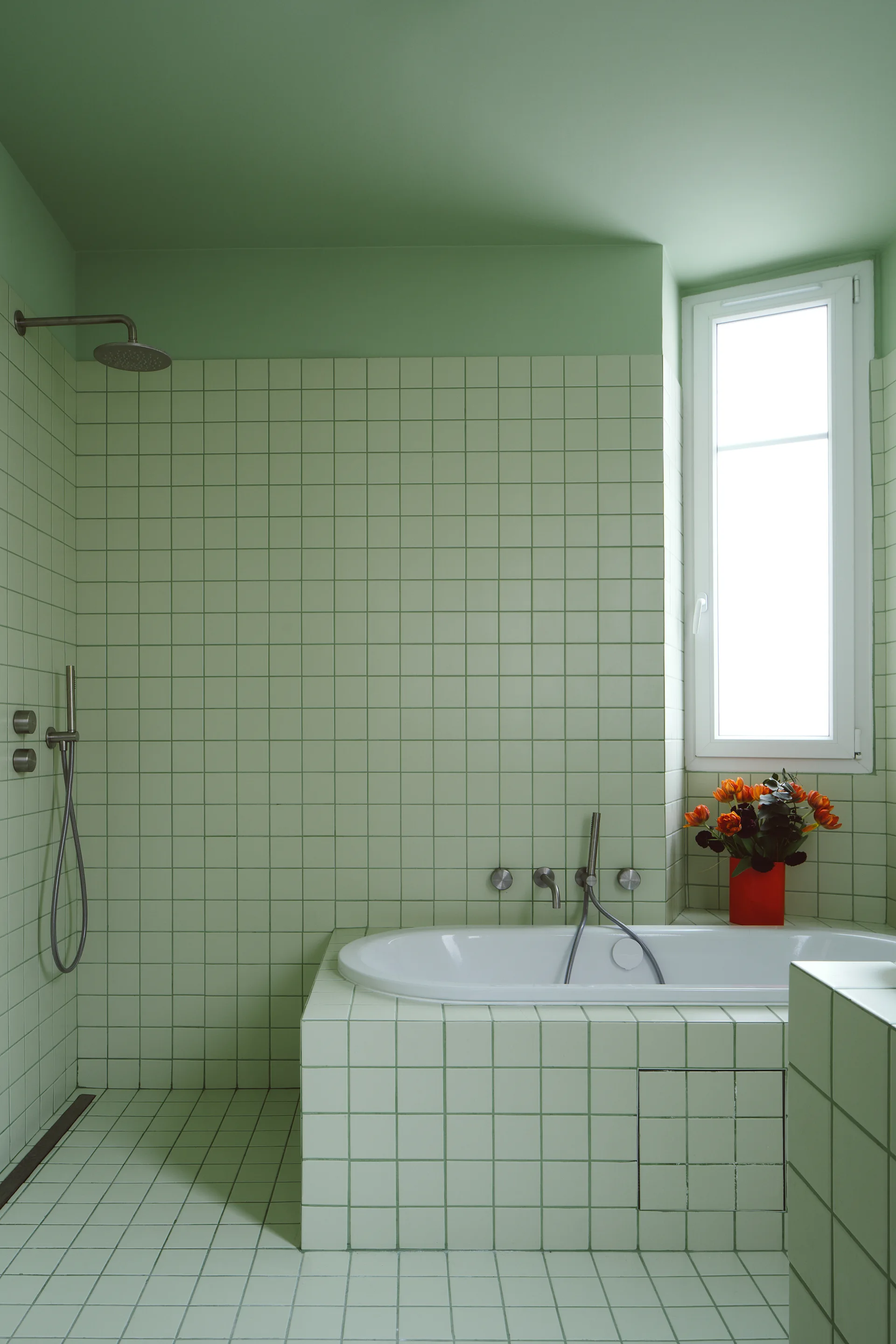 A minimalist bathroom features light green square tile on the walls, floor, and built-in bathtub next to a white-framed window.