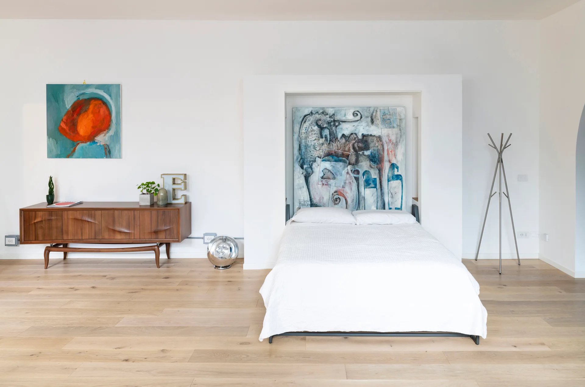 A bed with white linens is nestled in a wall alcove next to a mid-century modern wooden credenza and a metal coat rack on light wood floors.