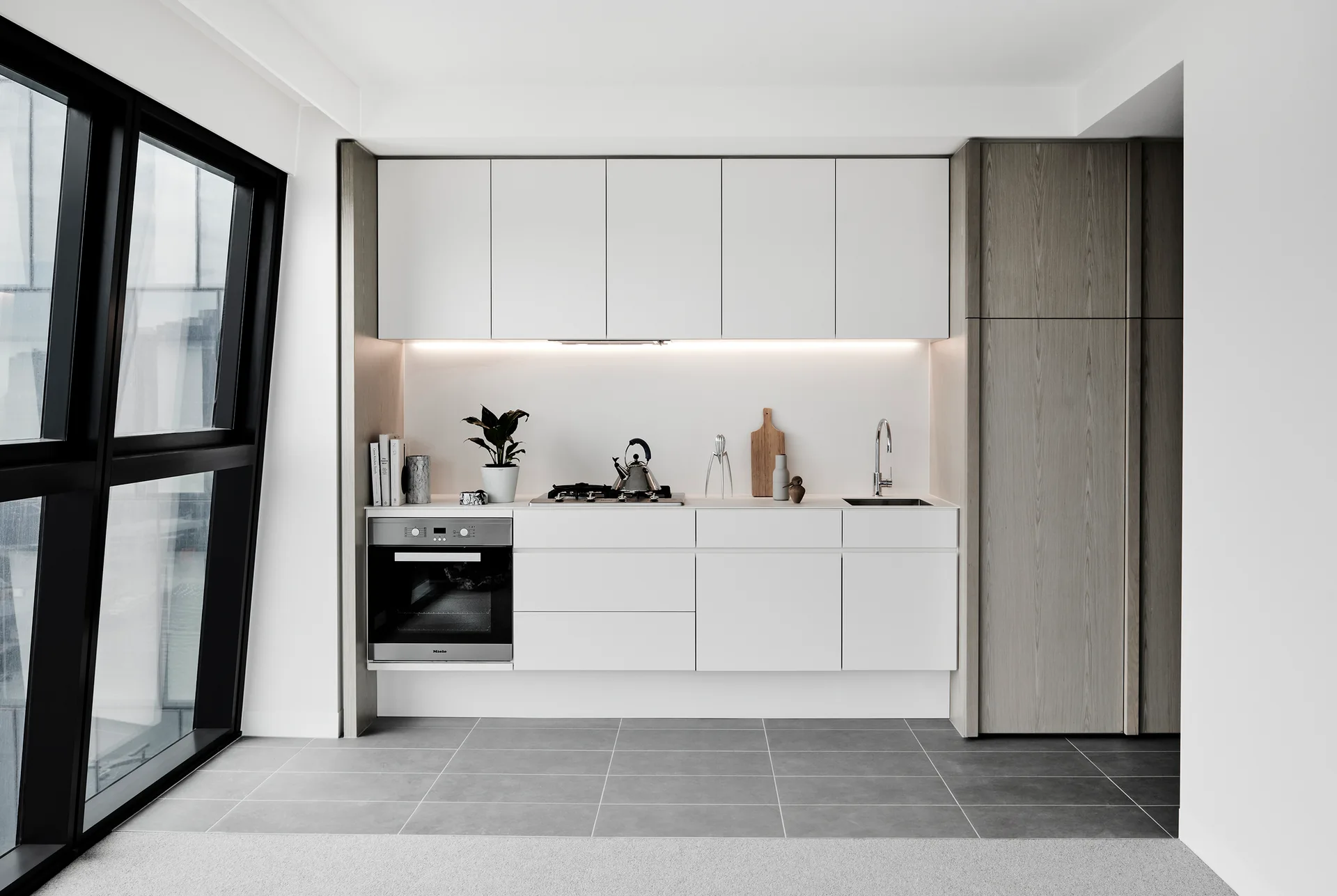 A minimalist white kitchen features flat-front floating cabinets, a wood-toned pantry, gray tiled flooring, and a large black-framed window.