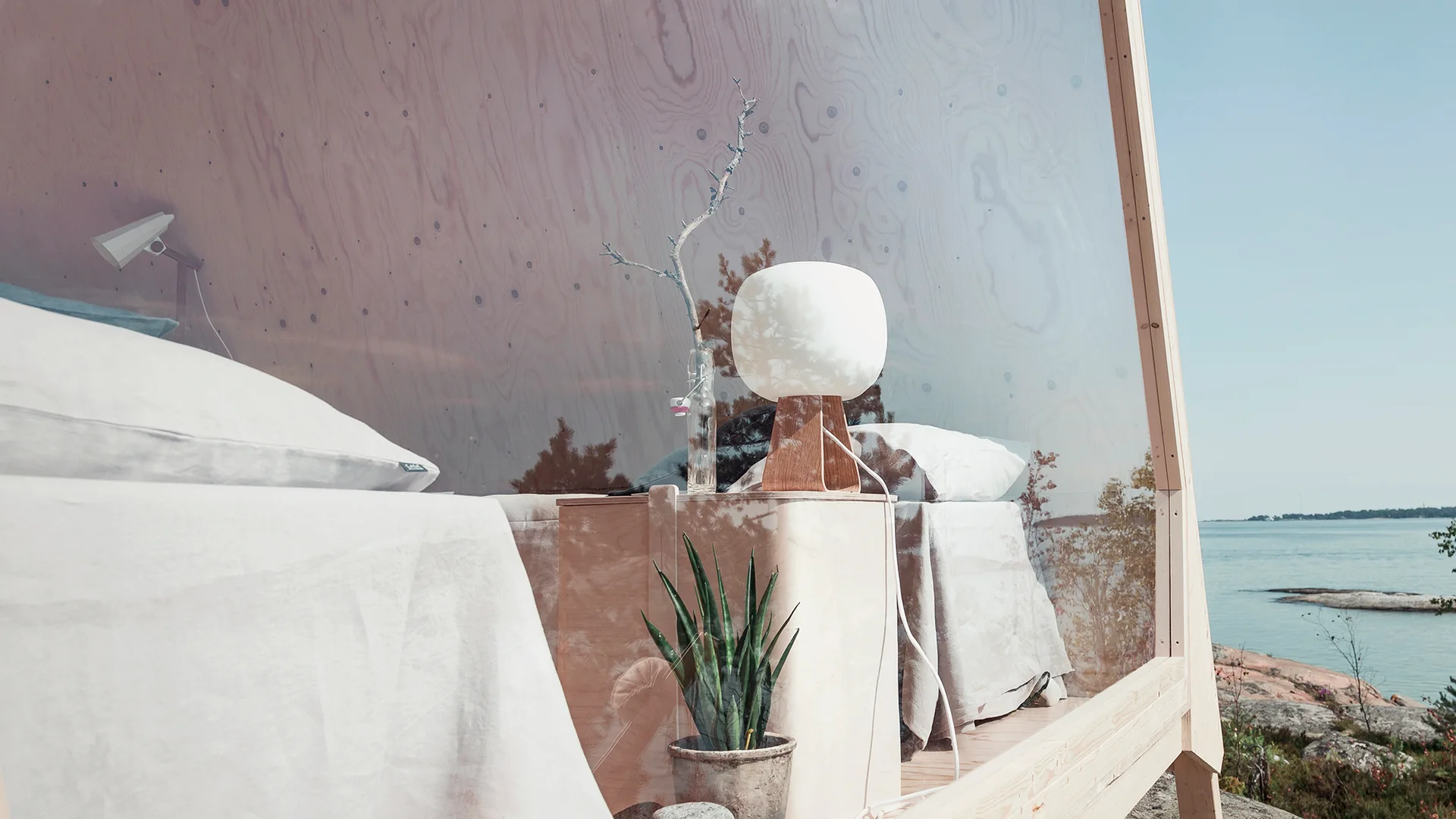 A minimalist bedroom with plywood wall paneling, white linen bedding, and a globe lamp features a glass wall overlooking a calm sea.