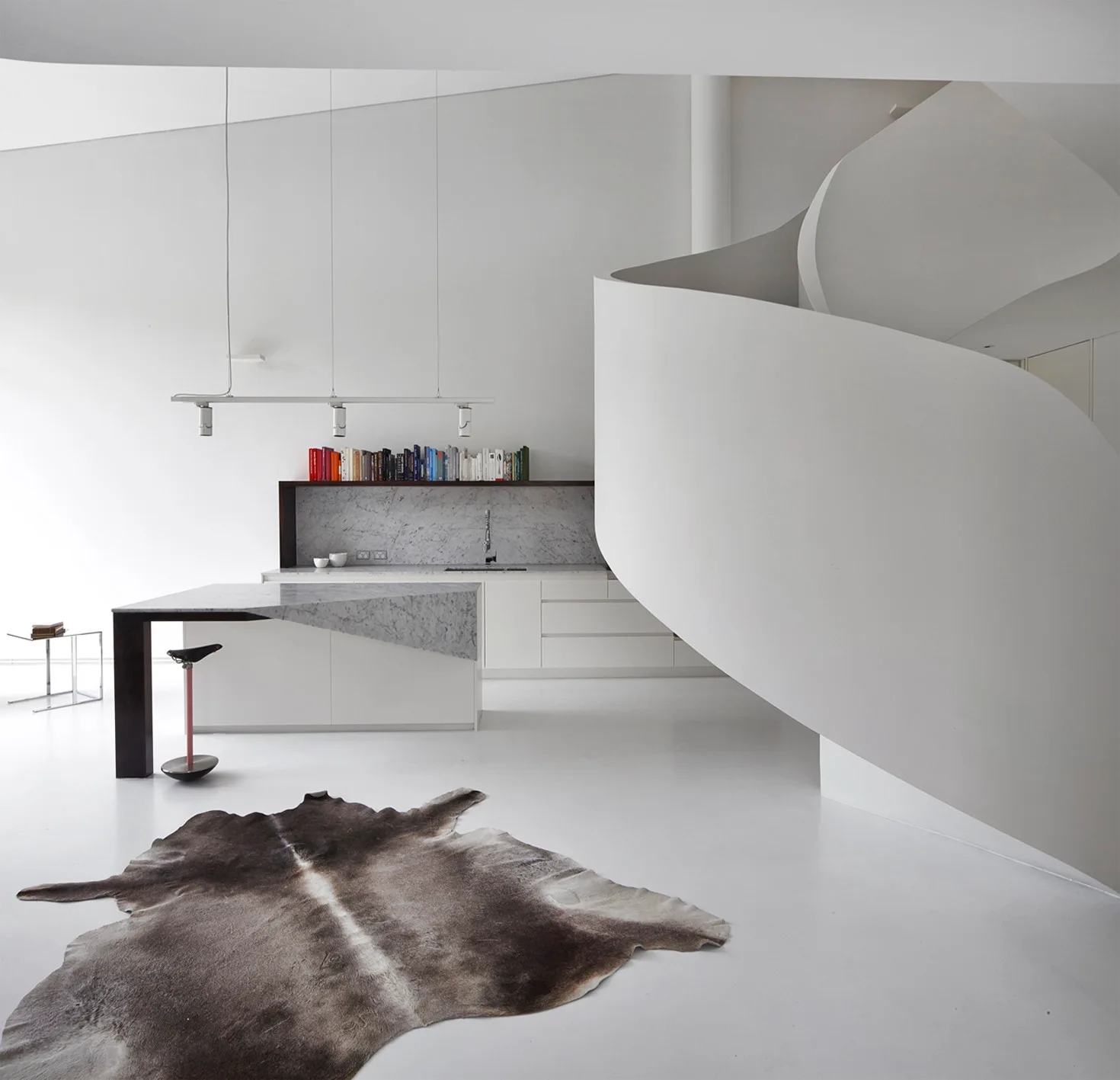 A minimalist white kitchen featuring a large curved white spiral staircase, a marble-topped island, and an animal hide rug on a seamless floor.