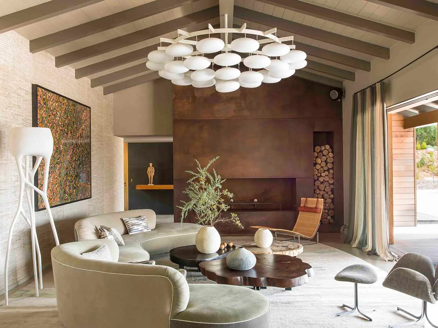 A living room features a curved sage green sofa, a prominent rusted metal fireplace, wooden ceiling beams, and a large white circular chandelier.