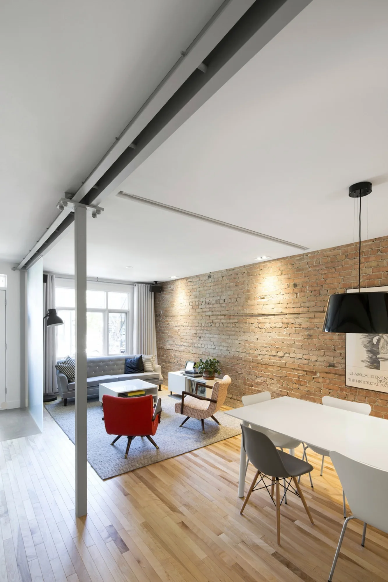 An open-plan living and dining area featuring an exposed brick wall, a gray sofa, a red armchair, and an industrial steel ceiling beam.