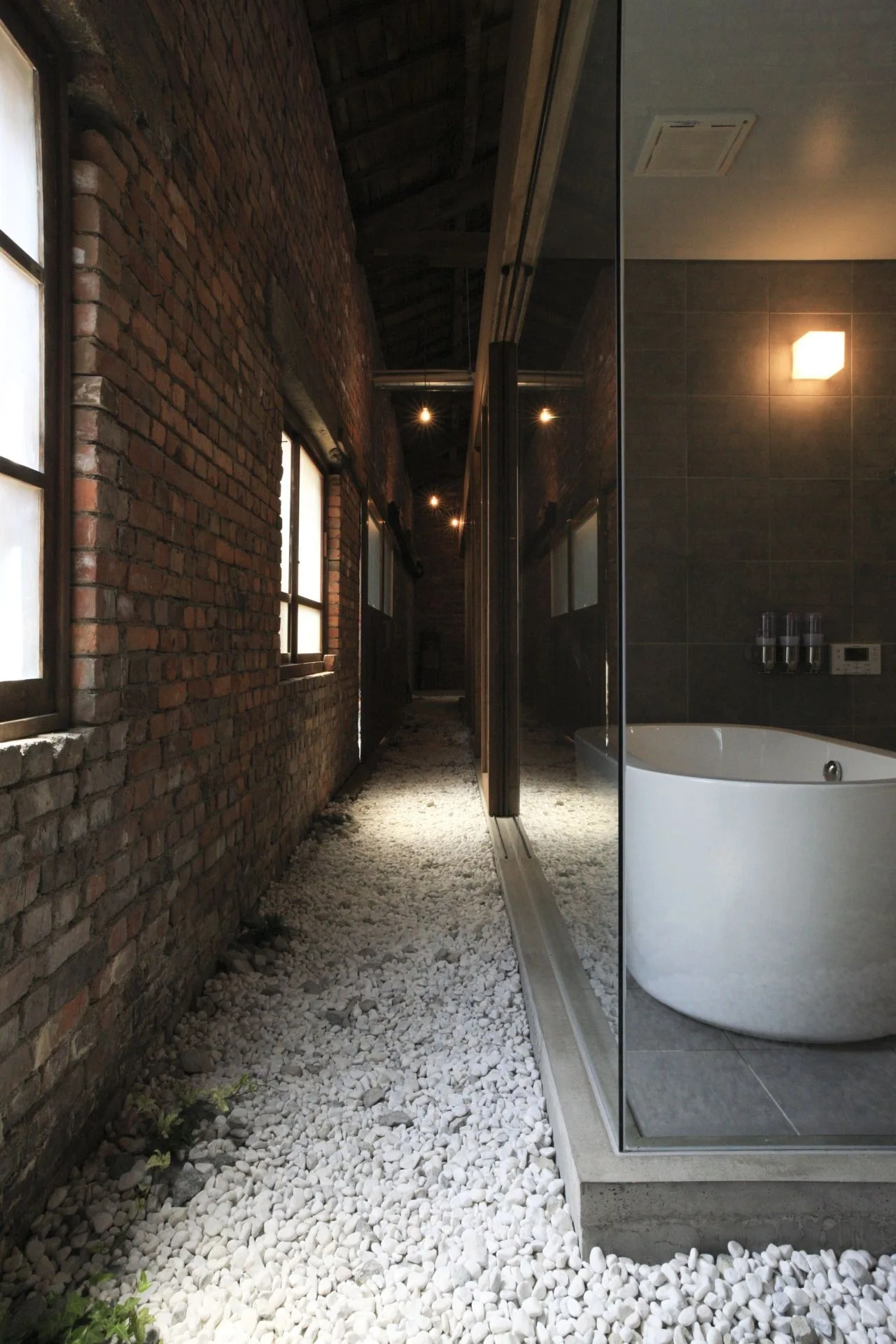 A modern white soaking tub sits behind a glass partition next to a corridor with an exposed red brick wall and white gravel floor.
