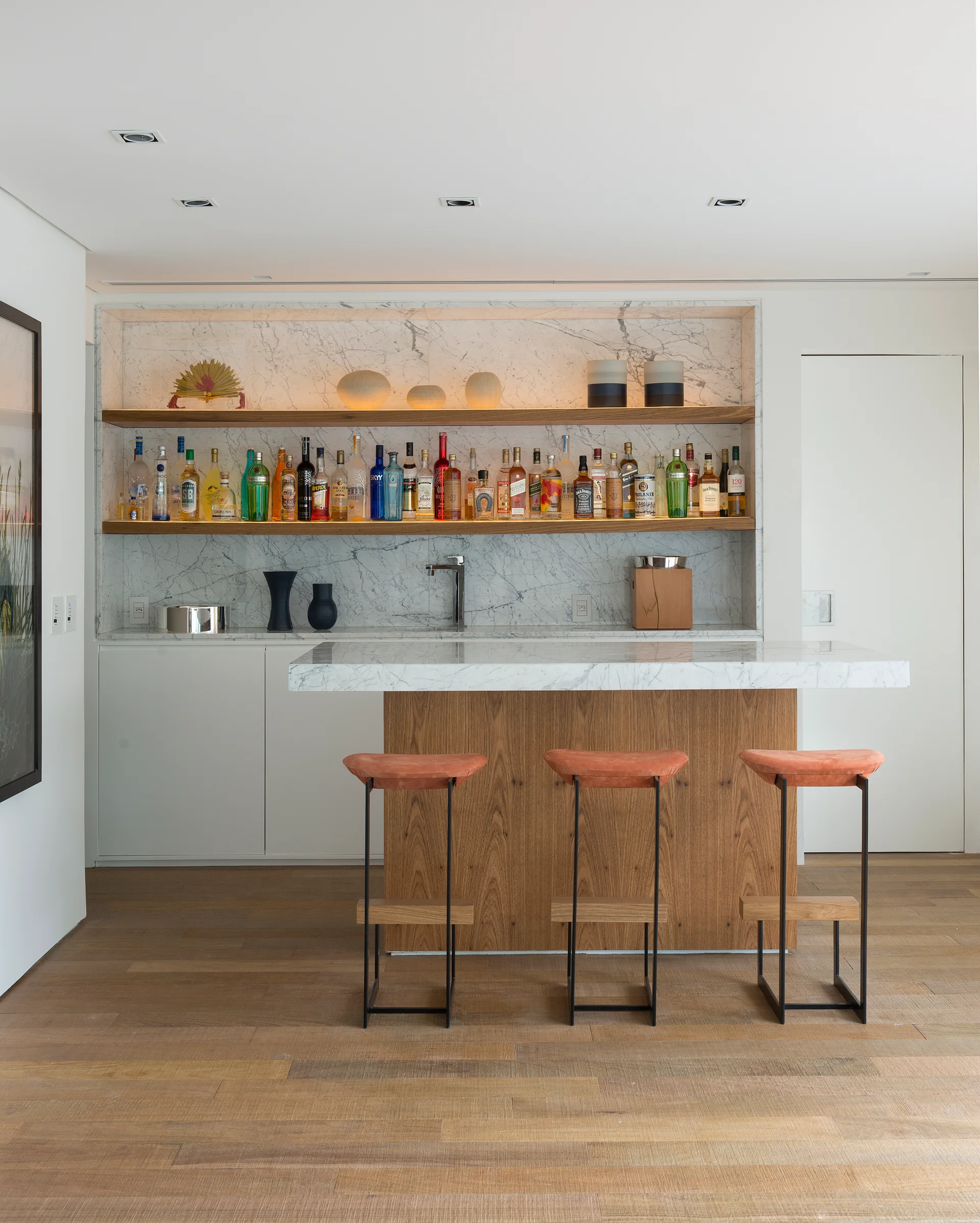 A modern home bar features a marble countertop, warm wood floating shelves with liquor bottles, and three orange-cushioned bar stools.