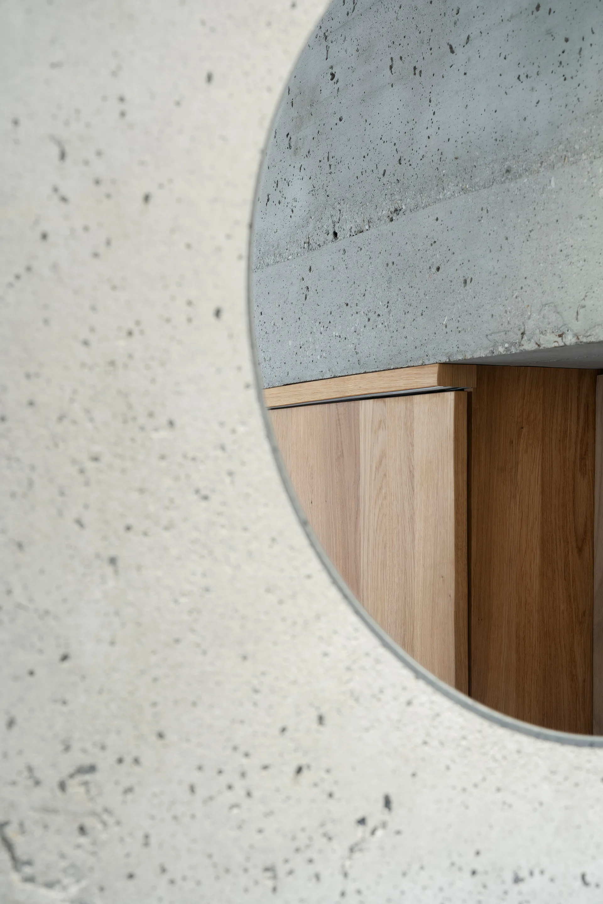 A circular opening in a textured gray concrete wall reveals a natural wood cabinet behind it.