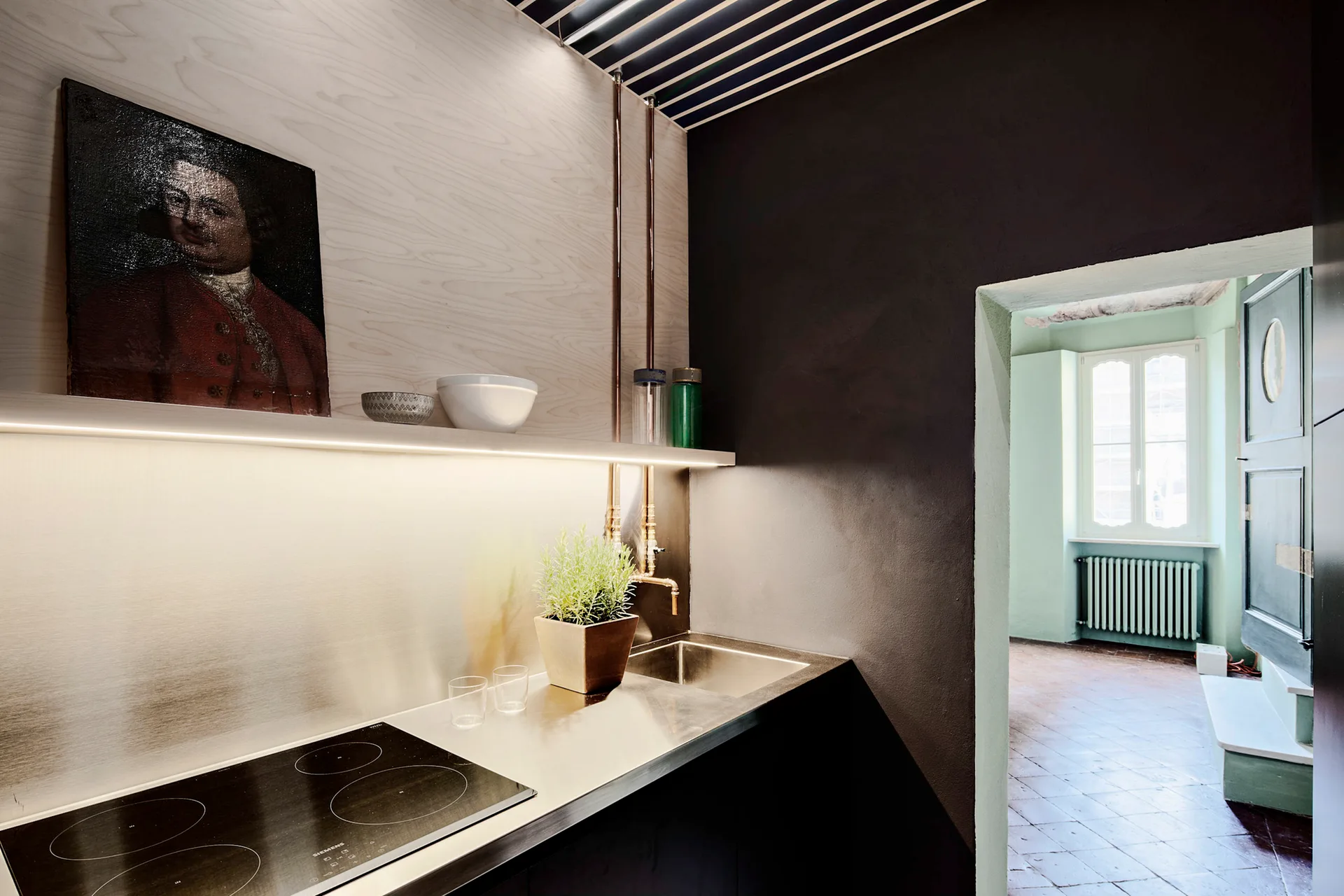 A modern kitchen space features a stainless steel countertop with an integrated sink and cooktop, light wood paneling, and an archway leading to another room.