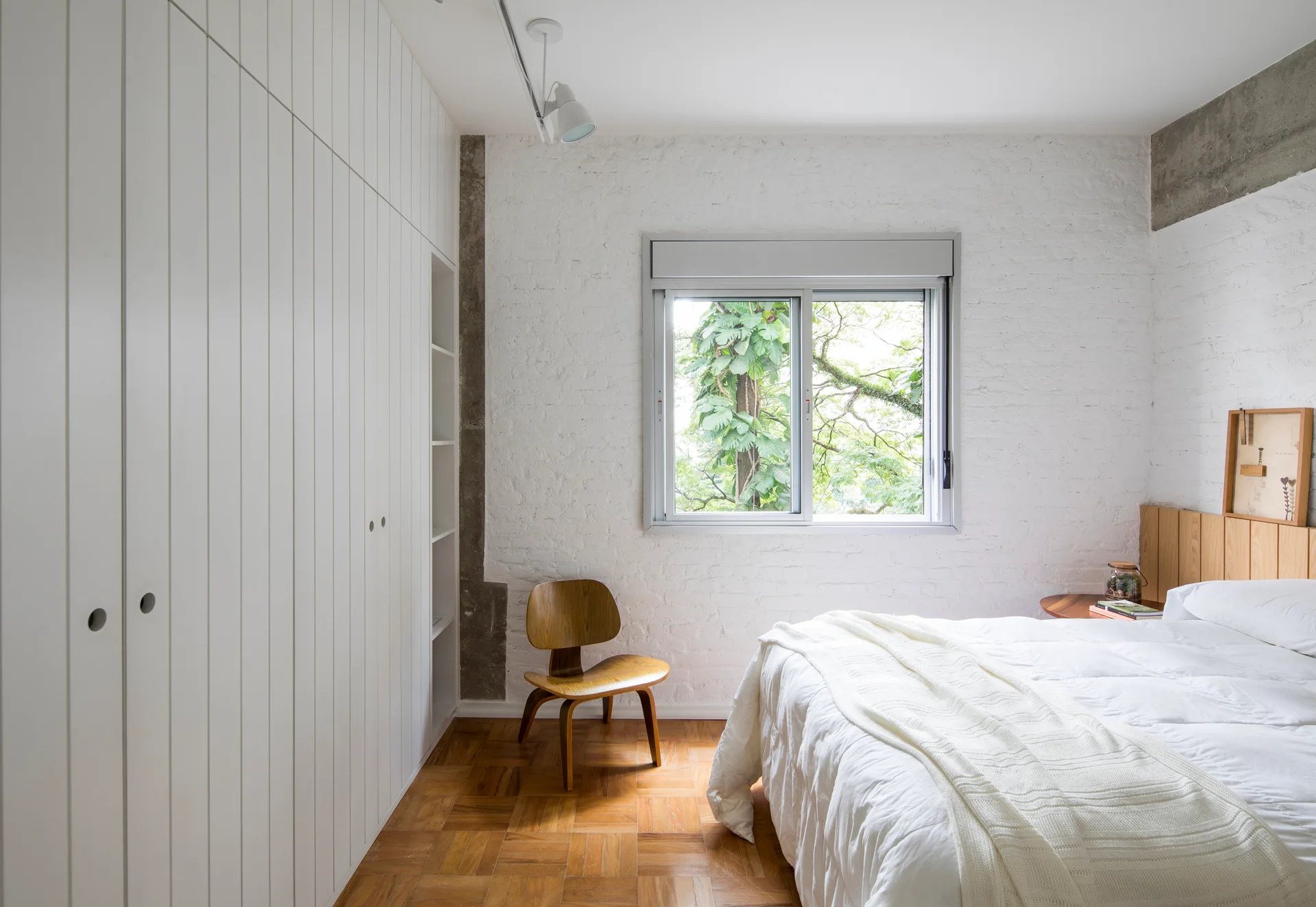 A minimalist bedroom featuring a white painted brick wall, built-in vertical paneled wardrobe, parquet wood floor, and a bed with white bedding.
