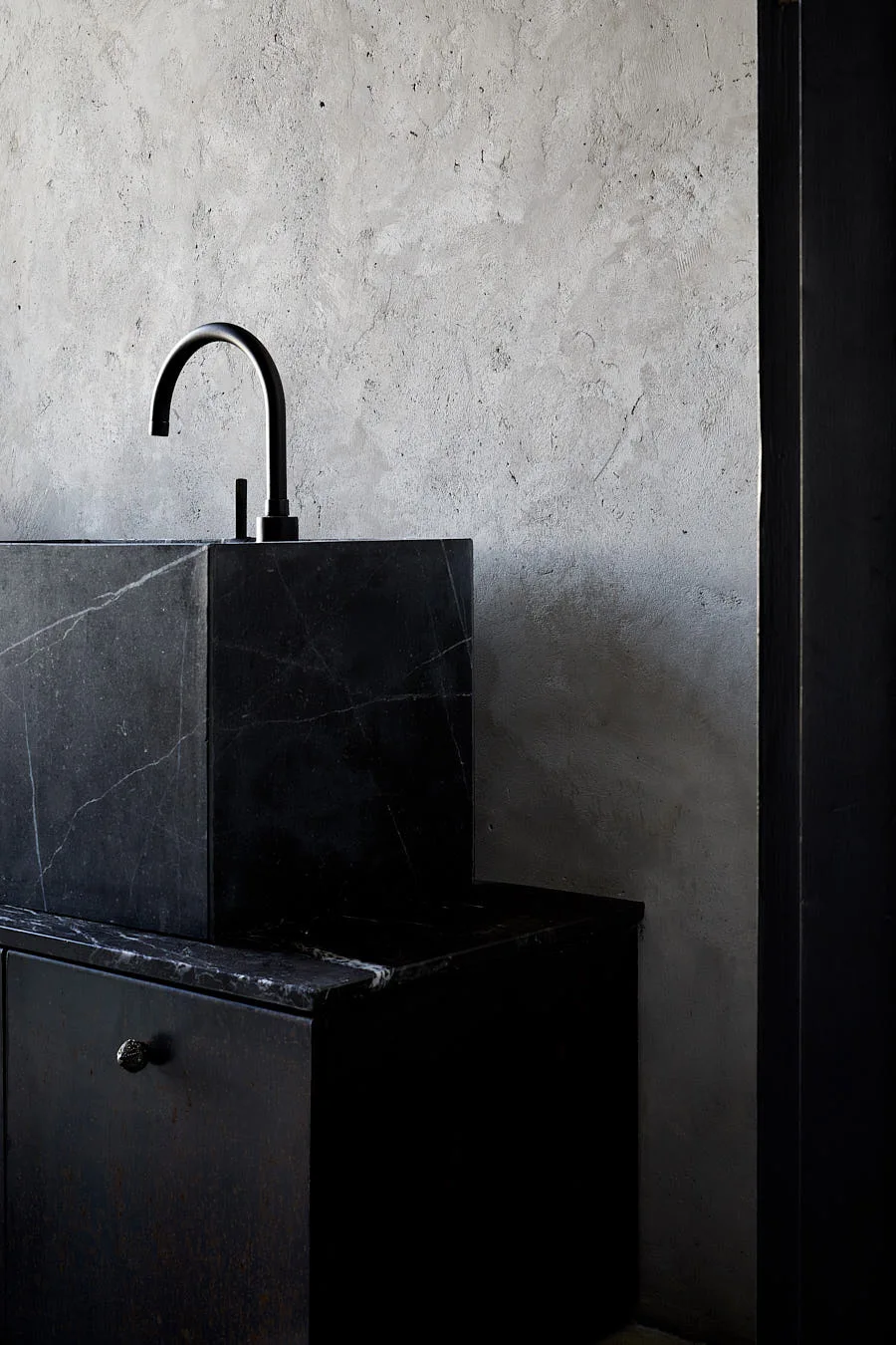 A cubic black marble vessel sink with a matte black arched faucet is set against a textured grey plaster wall.