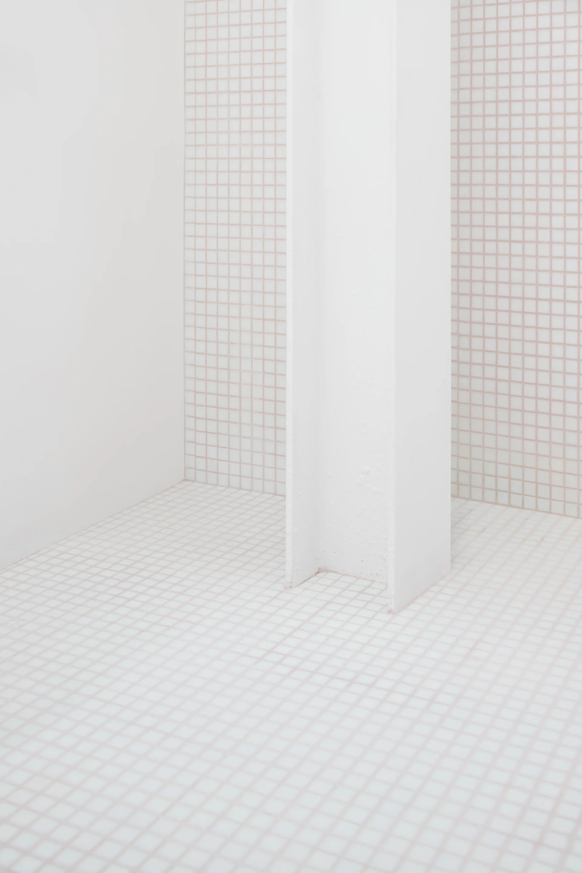 A minimalist room corner featuring floors and walls covered in small white square tiles with pink grout, with a white structural steel column.