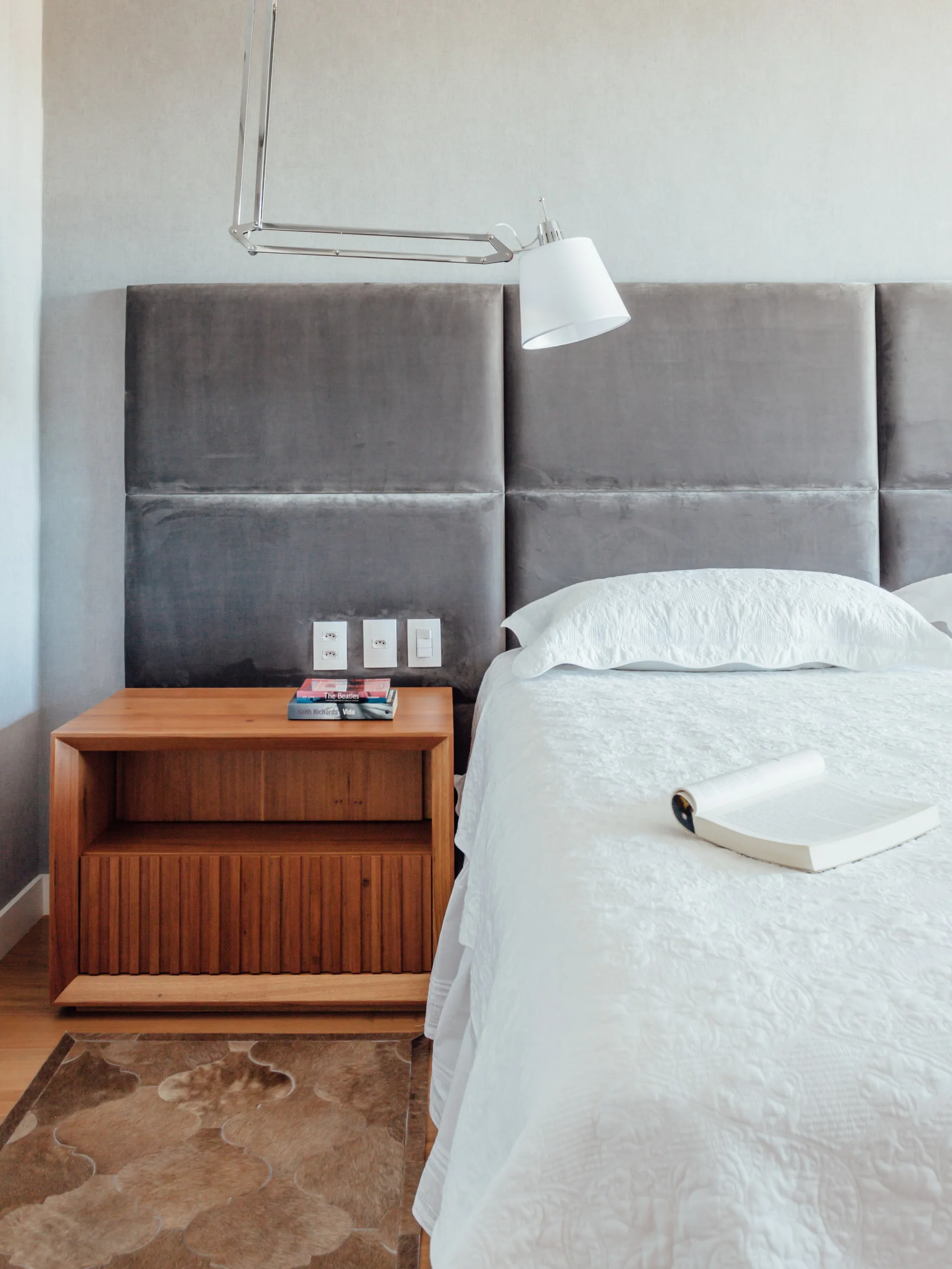 A bedroom corner features a grey velvet paneled headboard, a slatted wood nightstand, and a white articulated wall lamp above white bedding.