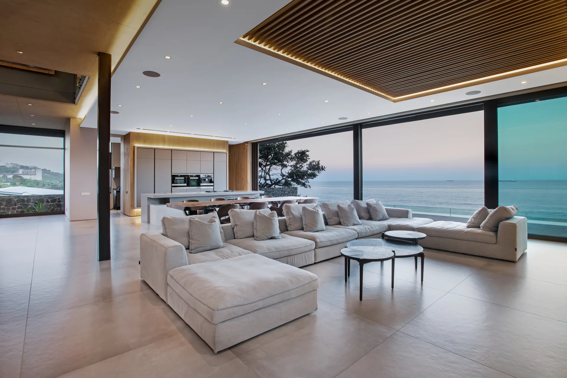 A large grey sectional sofa sits on a tiled floor in an open-plan living space with a wood-slat ceiling panel and floor-to-ceiling ocean views.