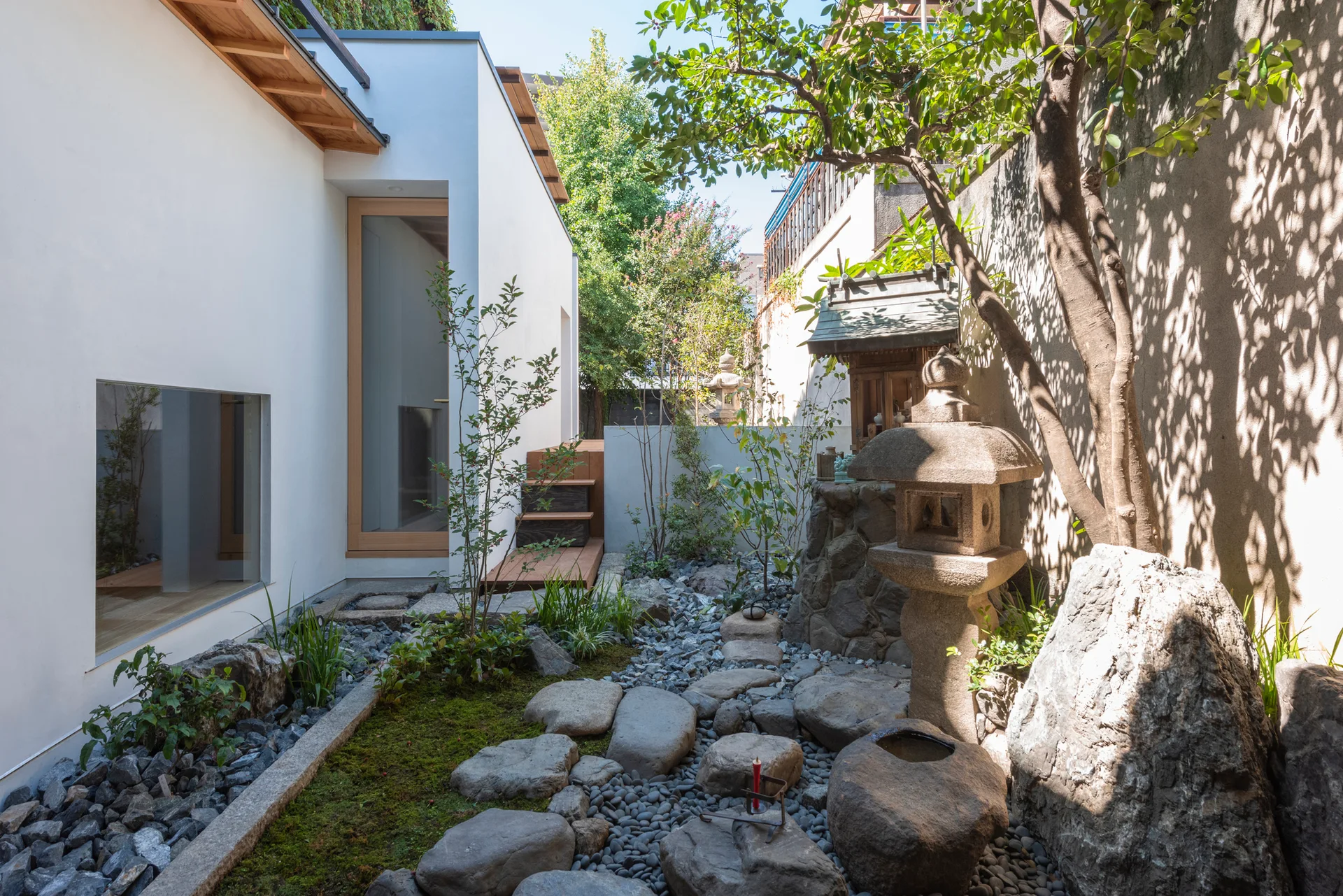 A Japanese-style courtyard garden featuring a stone lantern, stepping stones, and mossy ground alongside a modern white-walled building.