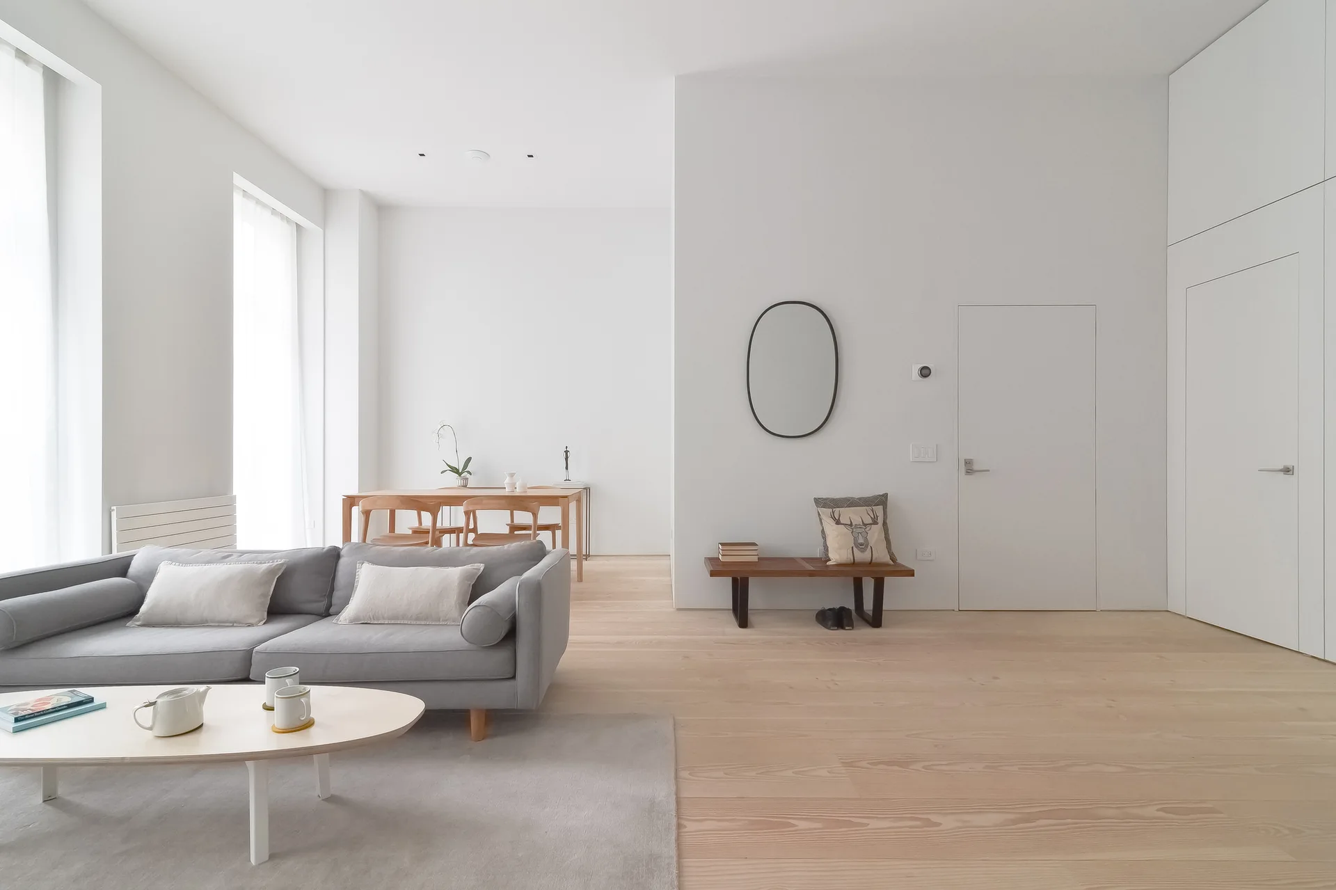 A minimalist living room features a grey sofa, an oval coffee table on a grey rug, and a wooden bench beneath an oval mirror on a white wall.