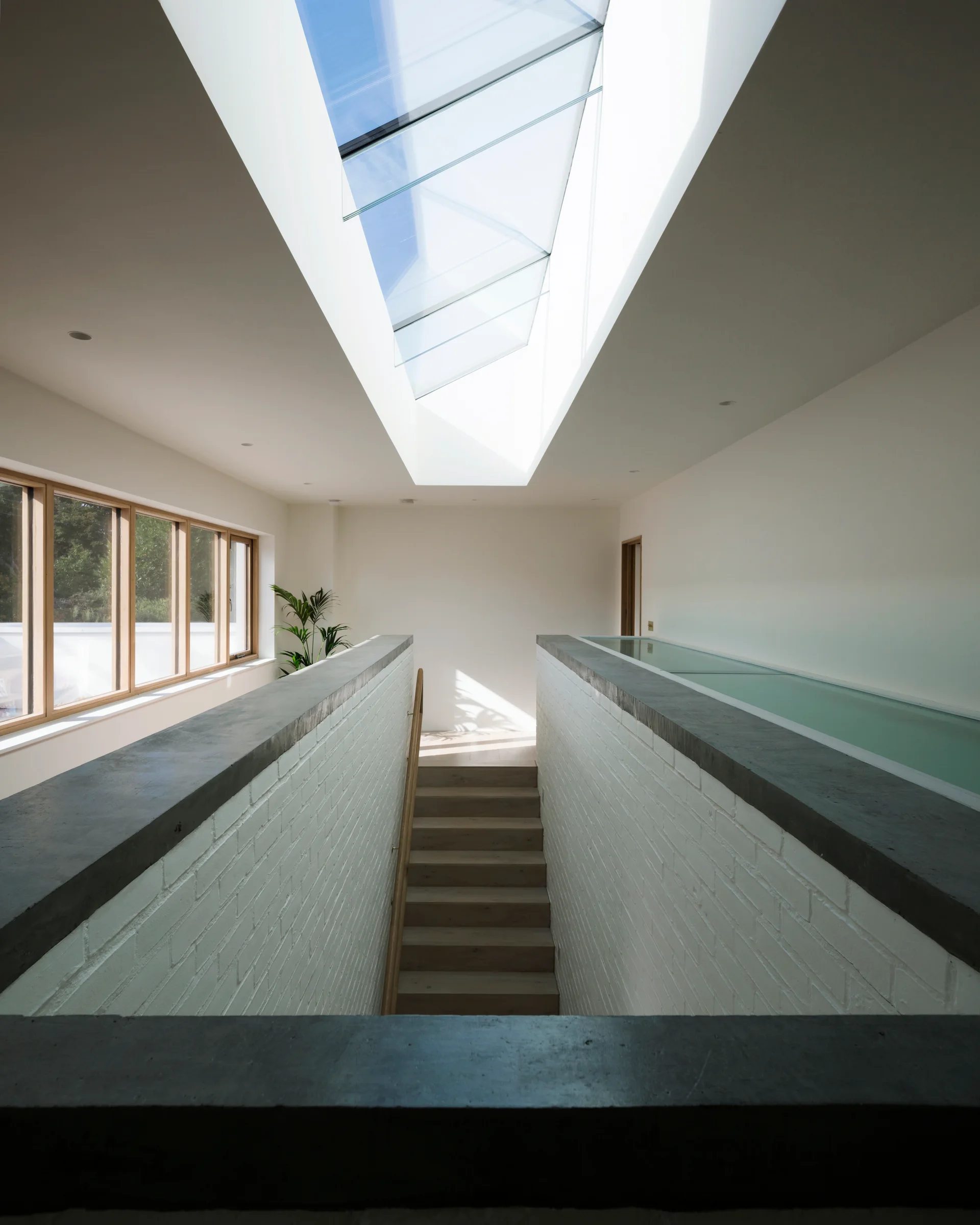 A wooden staircase descends between white painted brick walls illuminated by a large overhead skylight.