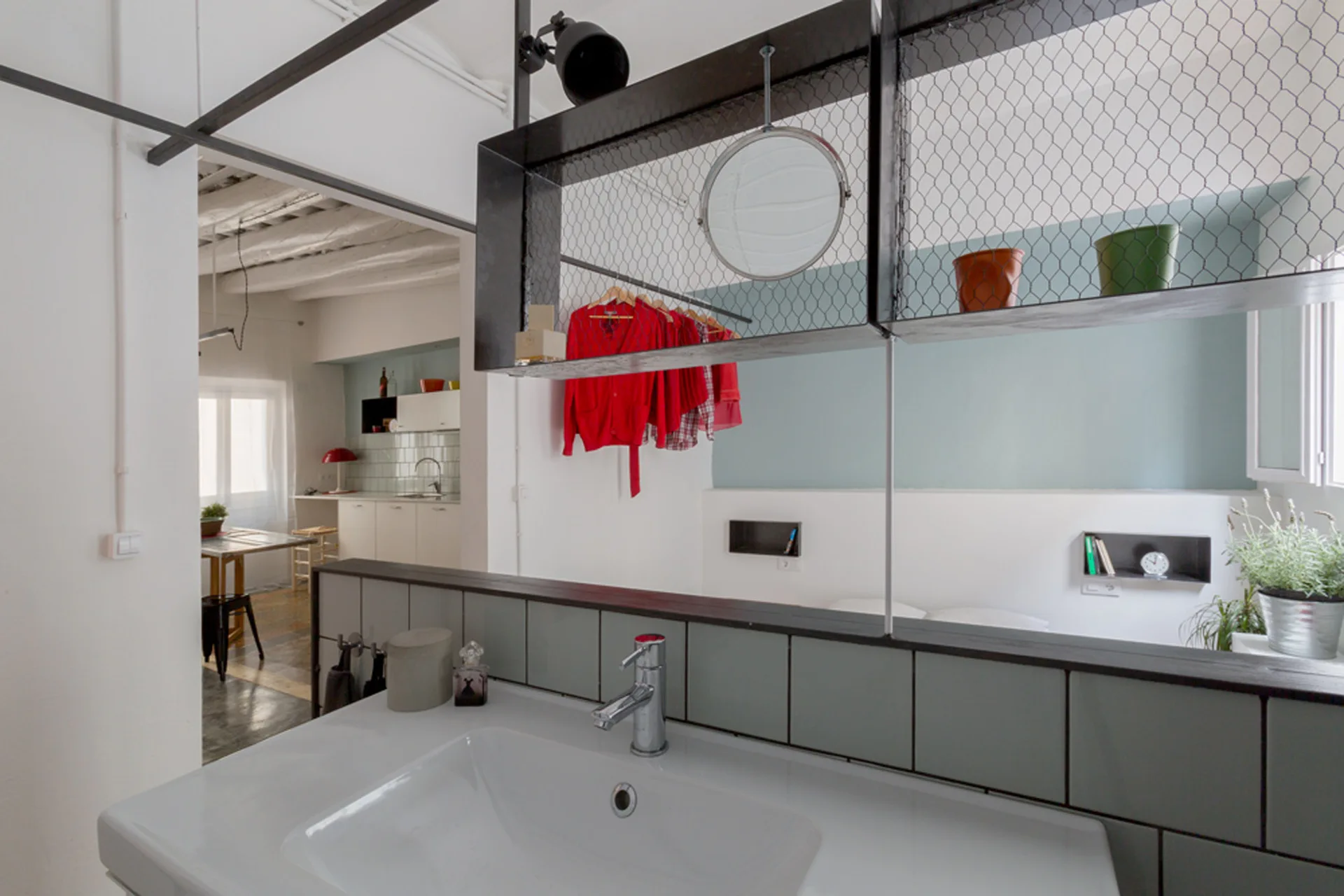 A bathroom vanity with a white ceramic sink features a tiled half-wall partition and a suspended black metal wire mesh storage shelf above it.