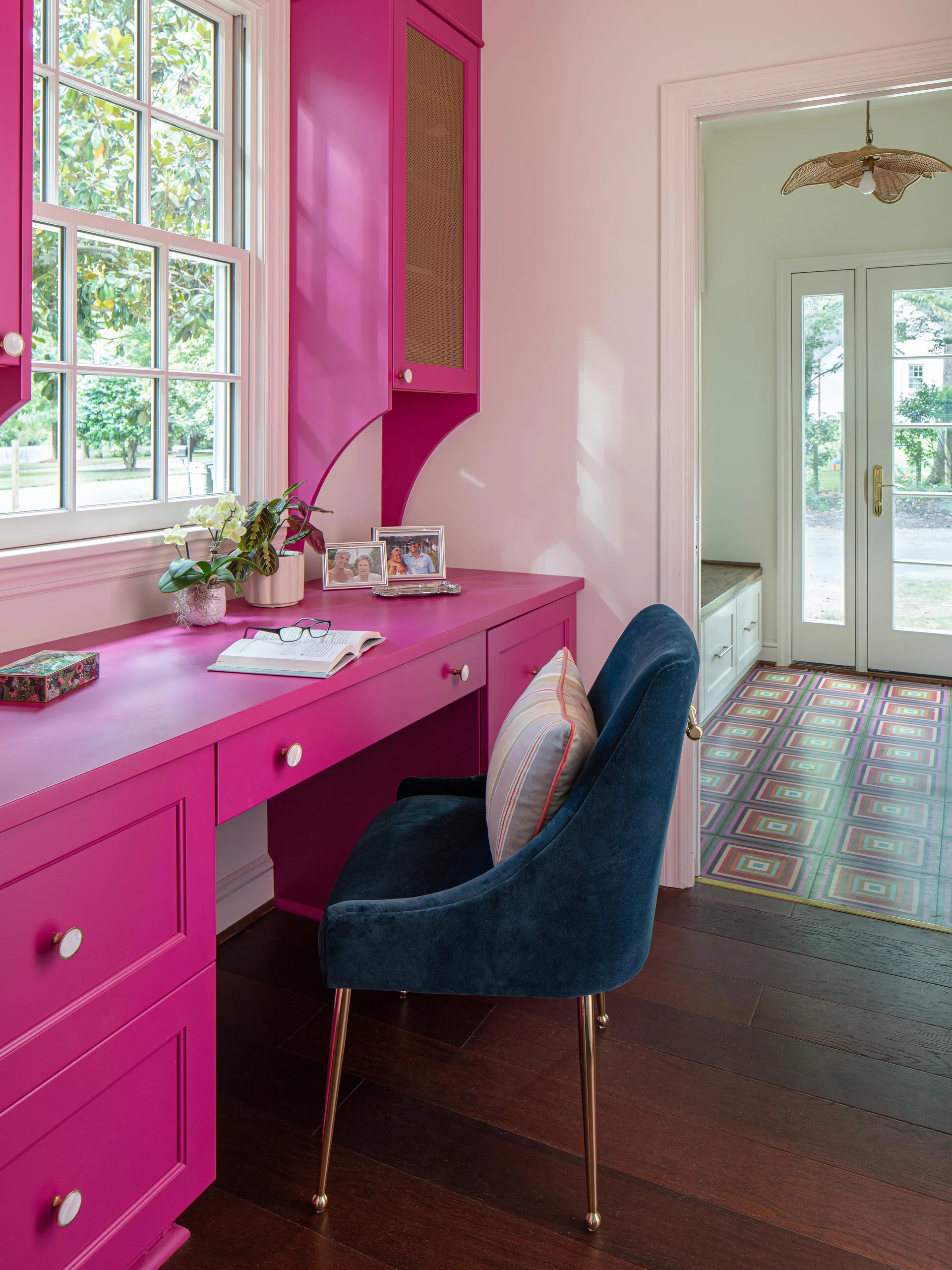 A bright fuchsia built-in desk with a navy blue velvet chair sits next to a paned window, showing a doorway leading to an entry hall.