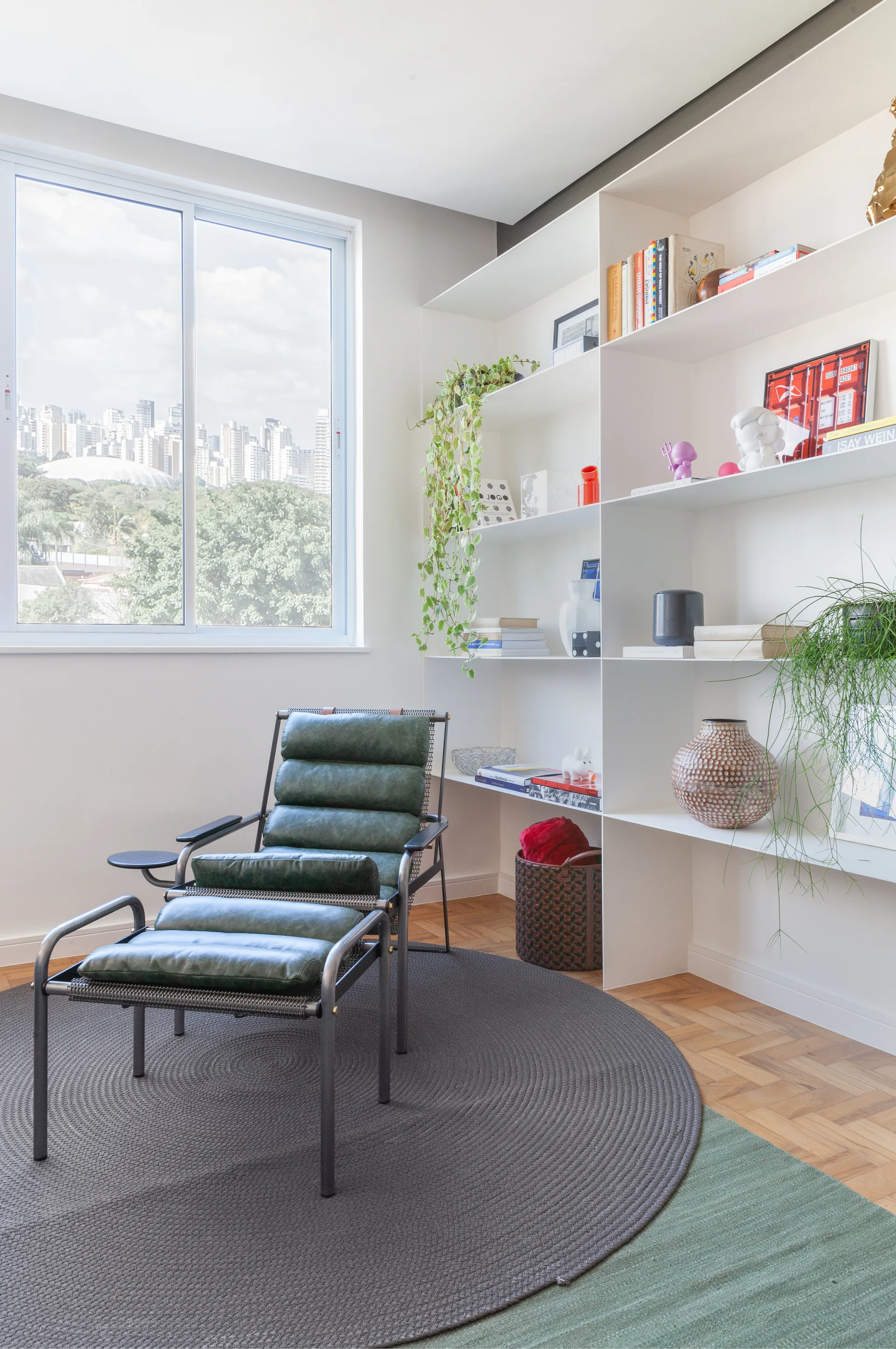 A green leather lounge chair with a footrest sits on overlapping rugs next to a tall white bookshelf and a window.