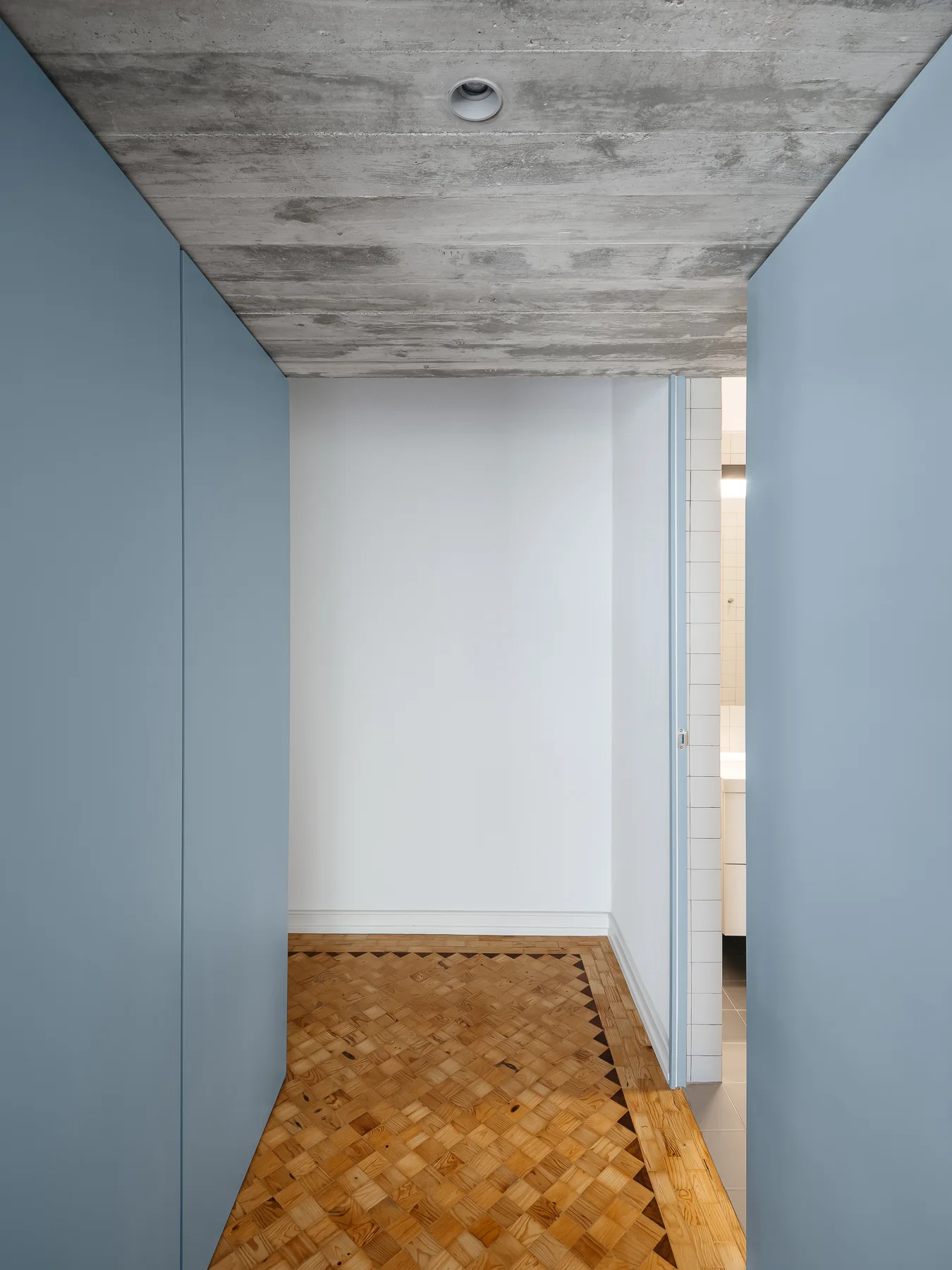 A narrow hallway features geometric parquet wood flooring, matte blue wall panels, and a raw concrete ceiling leading toward a white tiled bathroom.