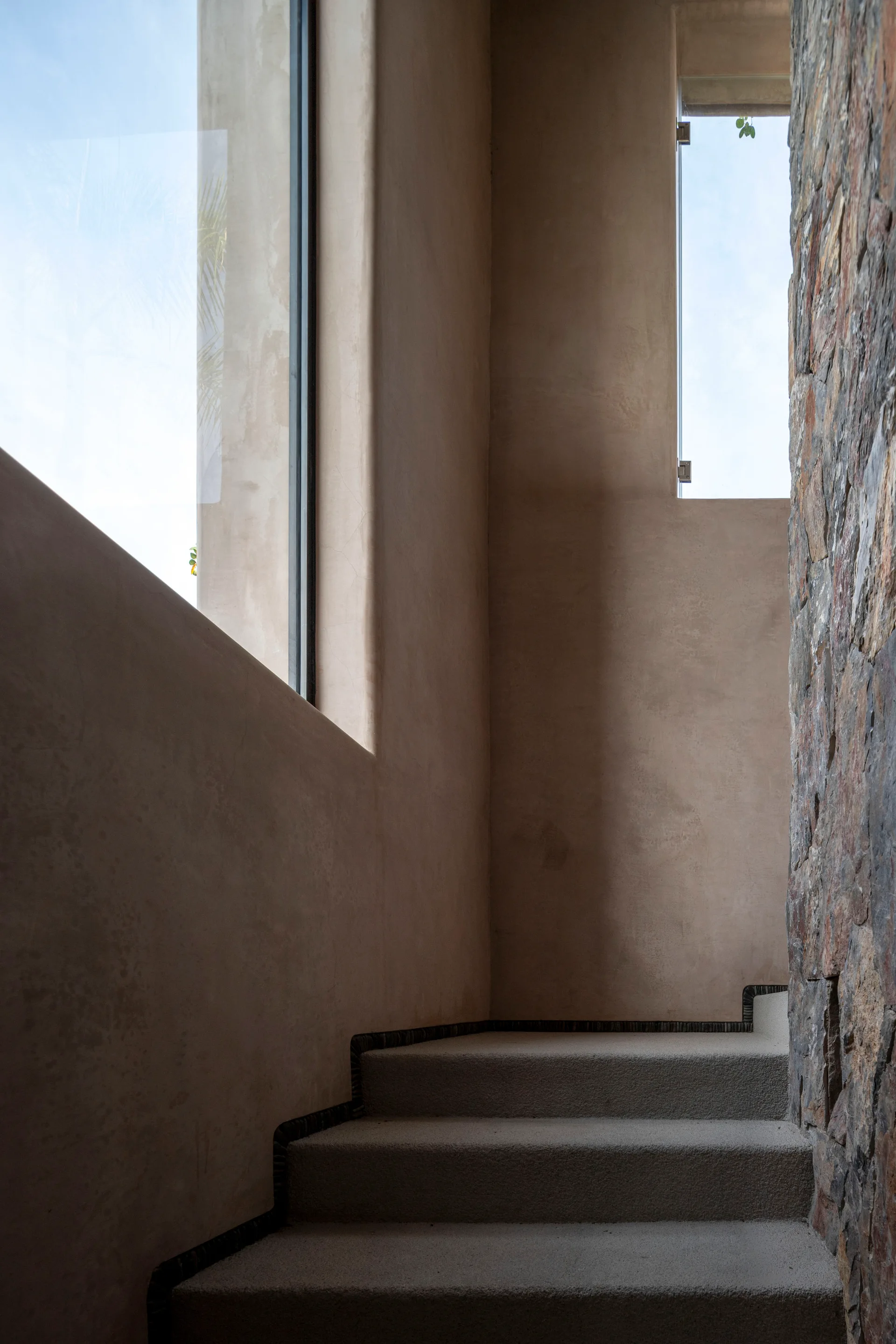 A cast concrete staircase is flanked by smooth tan plaster walls, a tall narrow window, and a rustic natural stone wall.