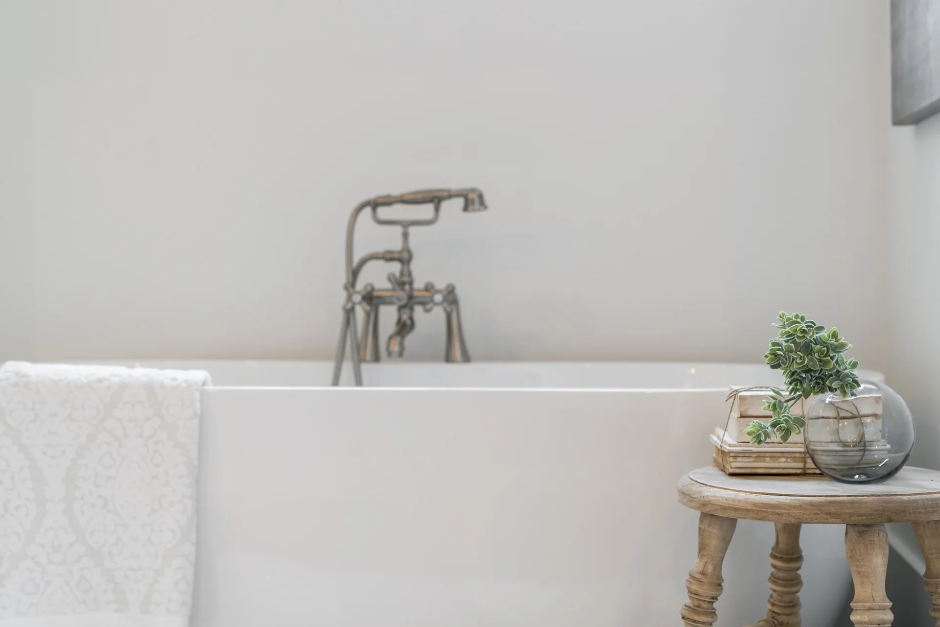 A white freestanding soaking tub features a metallic vintage-style faucet, a patterned towel, and a rustic carved wooden stool with books and a small plant.