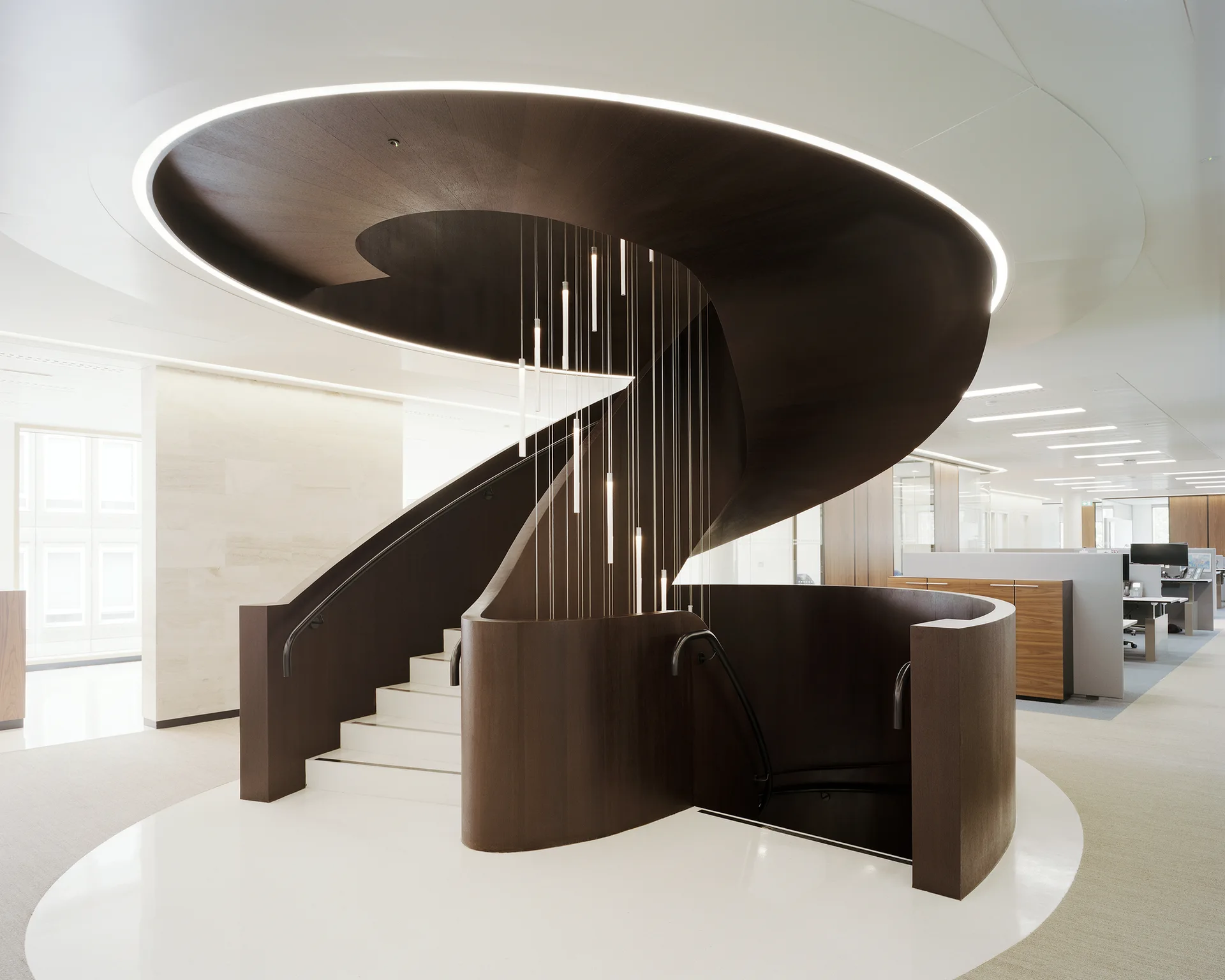 A dark wood spiral staircase with white steps and a cluster of vertical pendant lights sits on a white floor in a modern office.