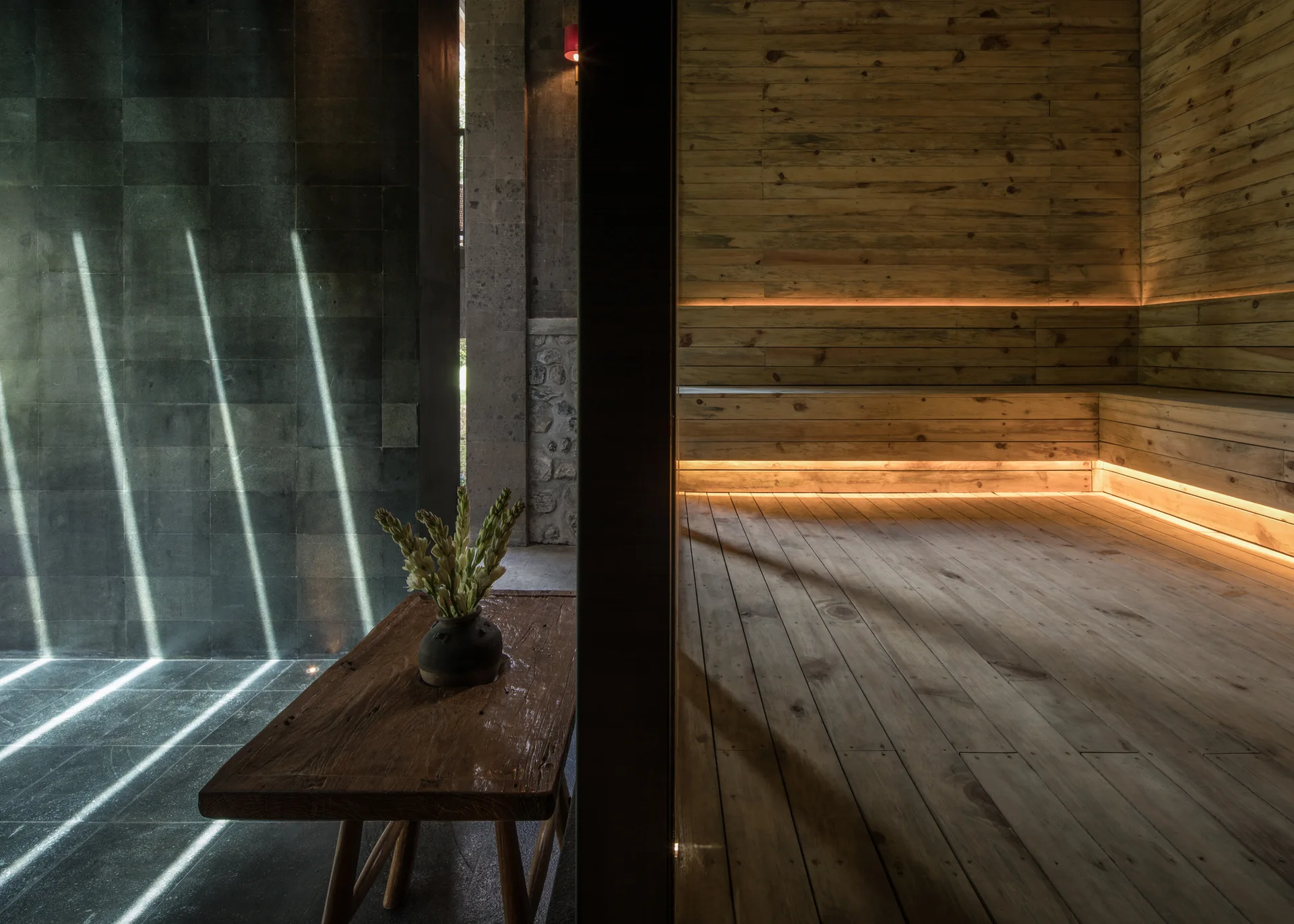 A split view of a warm wood-paneled sauna with built-in benches and integrated LED lighting next to a dark stone hallway with a rustic wood table.