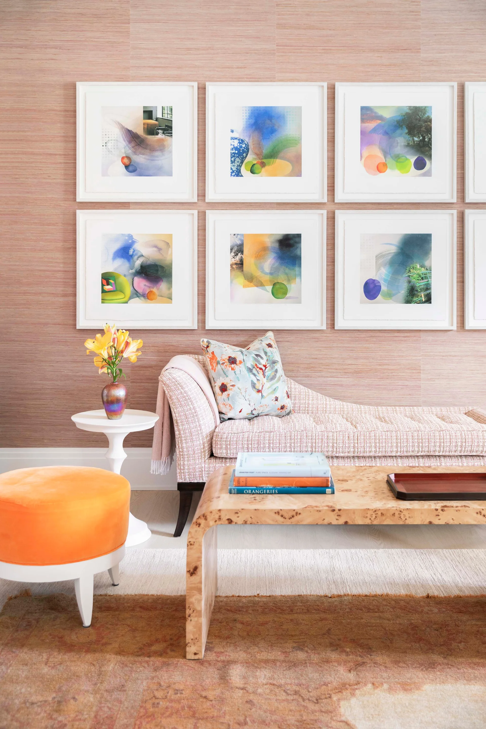 A pink grasscloth wall displays a six-print gallery grid above a patterned chaise lounge, a burl wood coffee table, and a round orange ottoman.