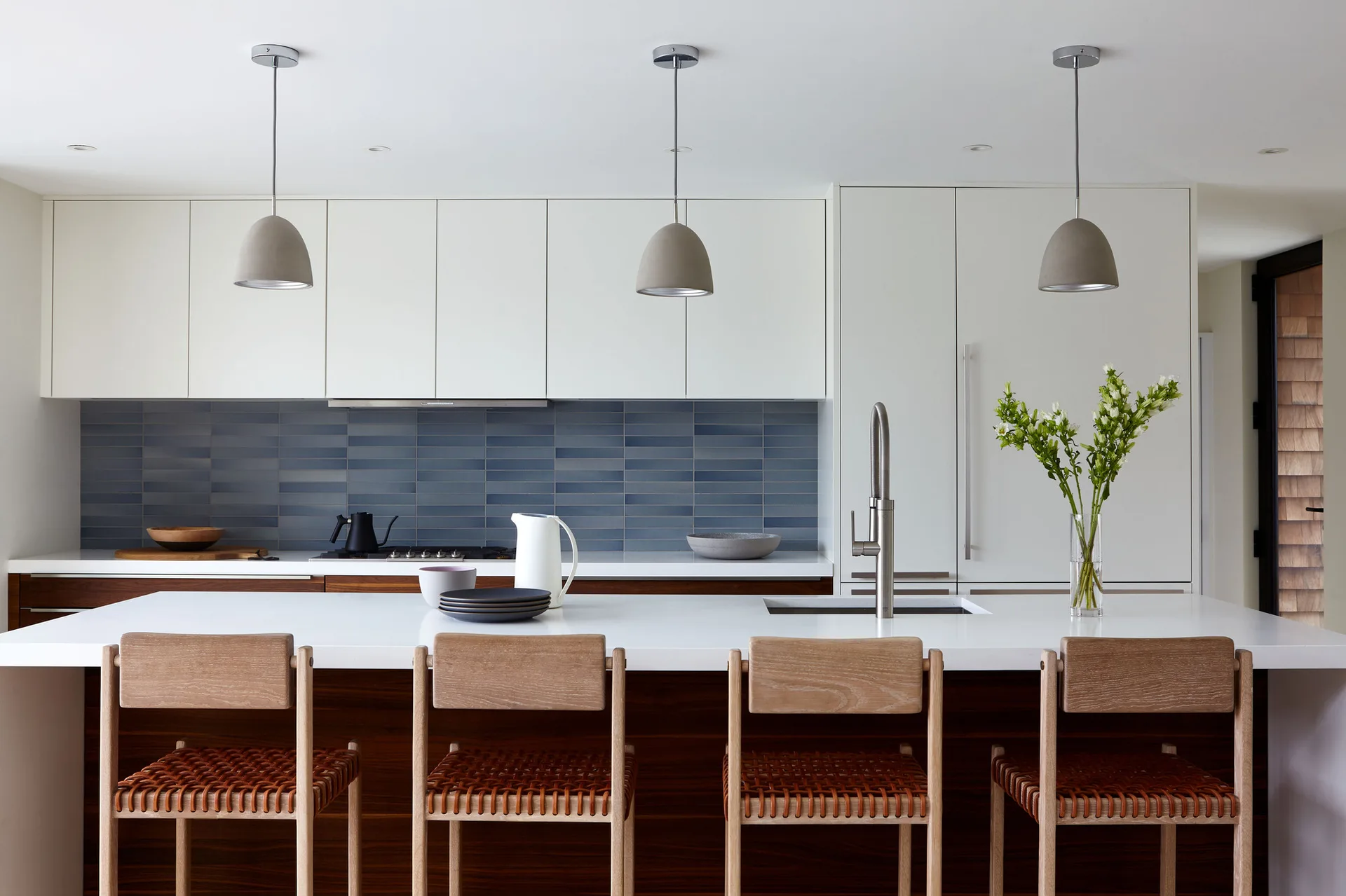 A modern kitchen features a white island with four woven leather stools, gray pendant lights, white upper cabinets, and a blue tiled backsplash.