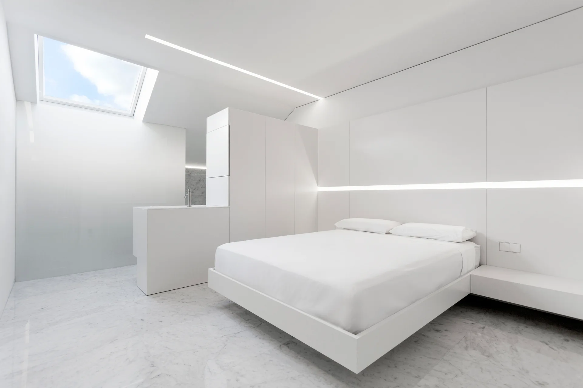 A monochromatic white bedroom features a minimalist platform bed, integrated linear wall lighting, a sloped ceiling with a skylight, and marble flooring.