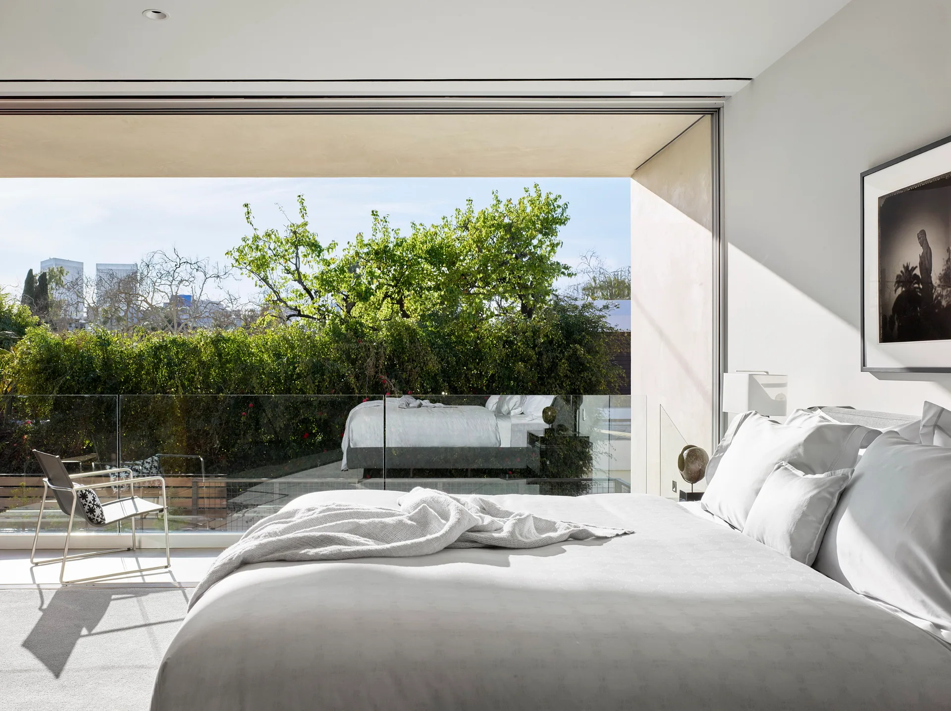 A bright minimalist bedroom features a large bed with white linens next to a sliding glass door opening to a sunlit balcony with green trees.