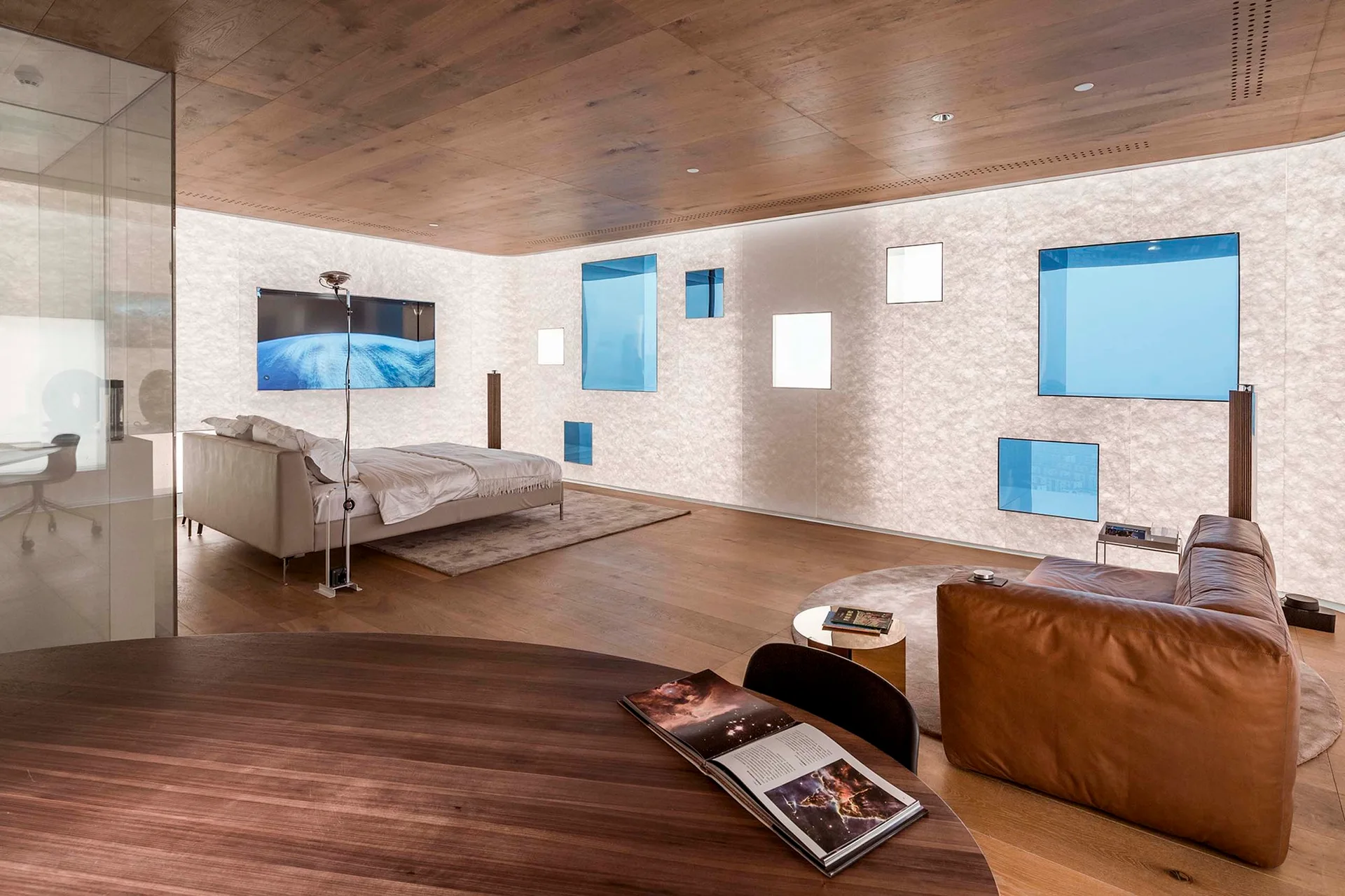 A minimalist bedroom features wood paneled floors and ceiling, a low grey bed, a brown leather sofa, and a glowing backlit wall with blue glass panels.