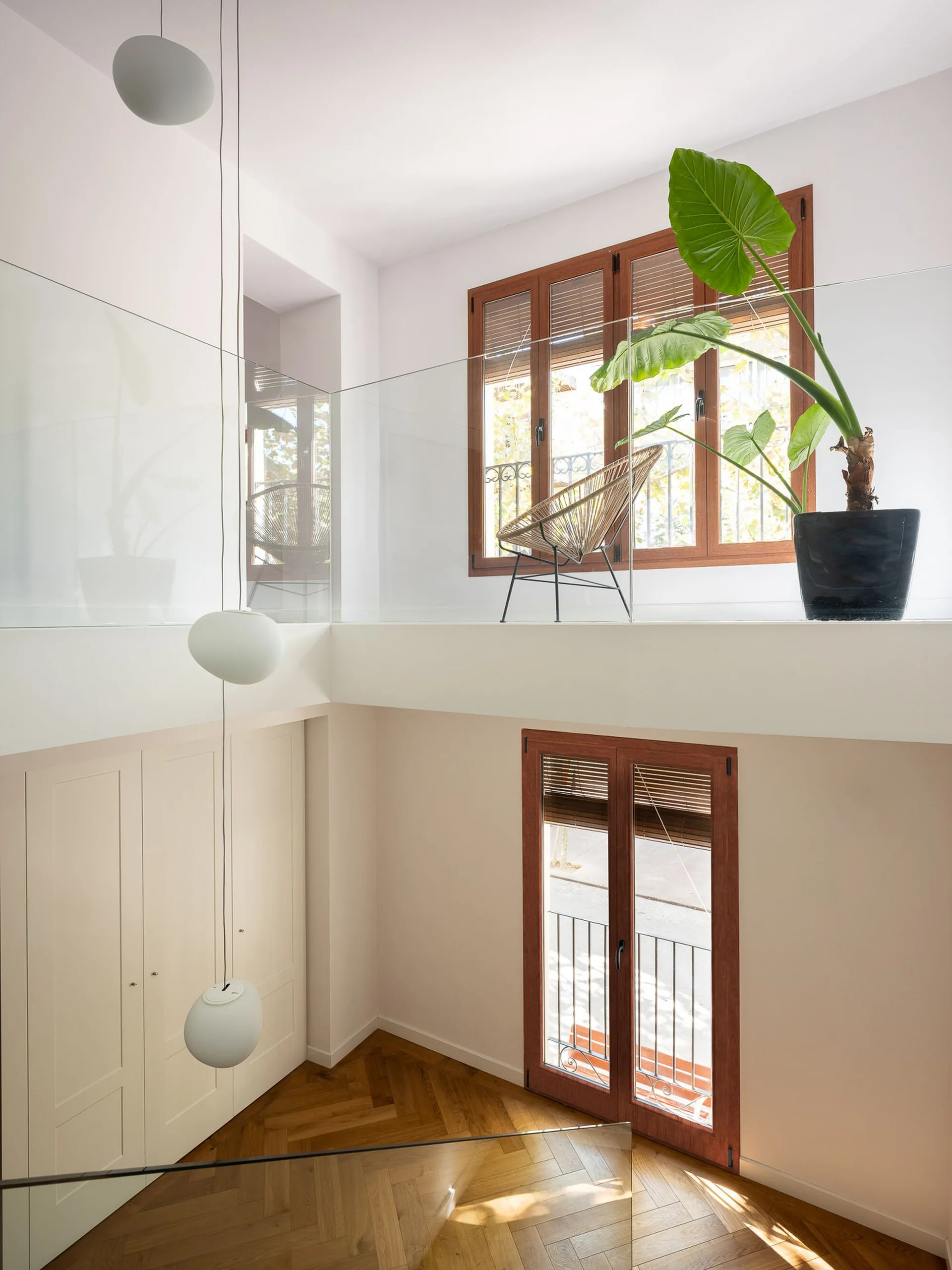 A modern mezzanine landing features a glass railing, herringbone wood flooring, wood-framed windows, and hanging globe pendant lights.