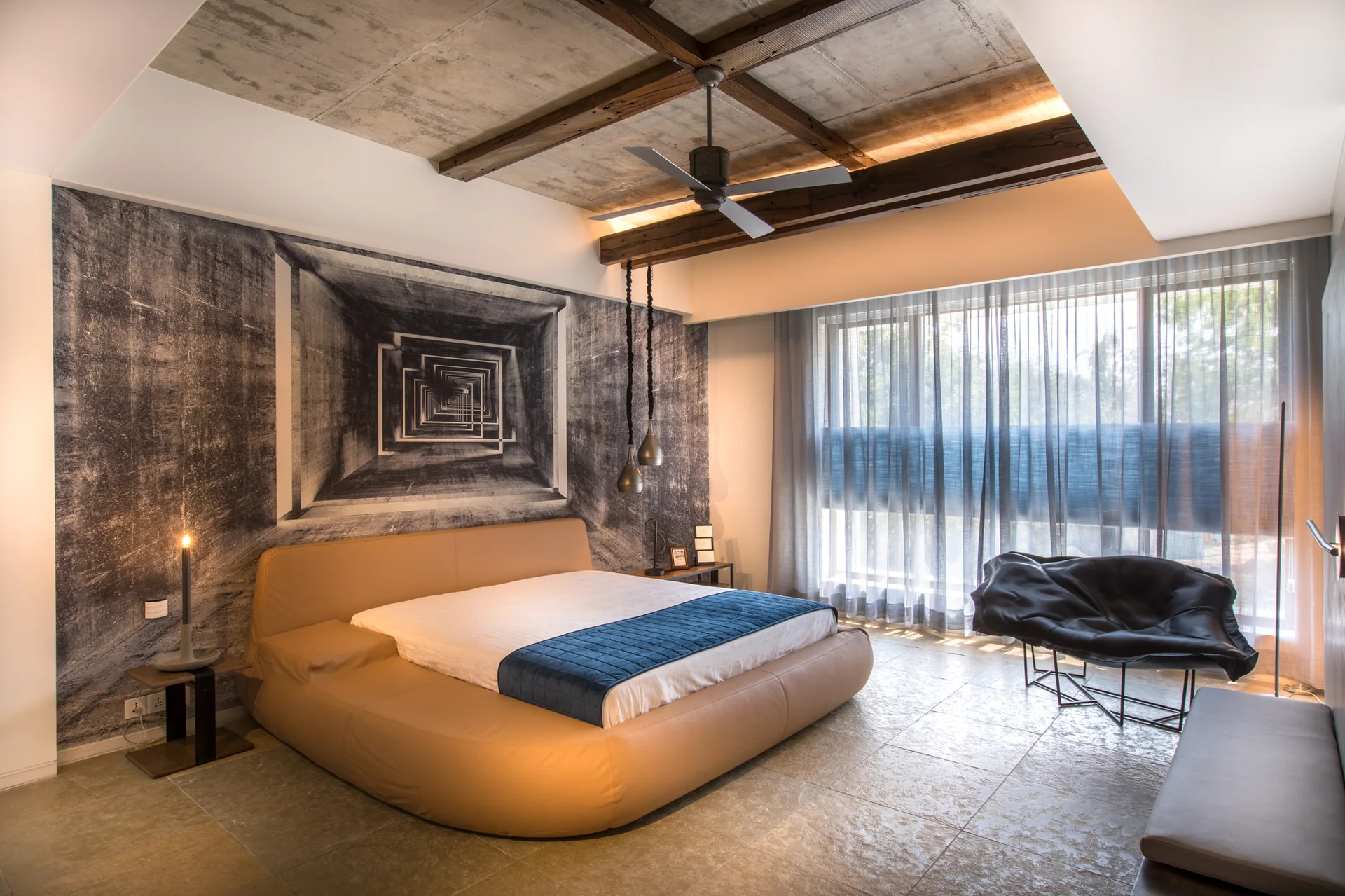 A contemporary bedroom features a low tan platform bed set against a gray geometric mural wall under an exposed concrete ceiling with timber beams.