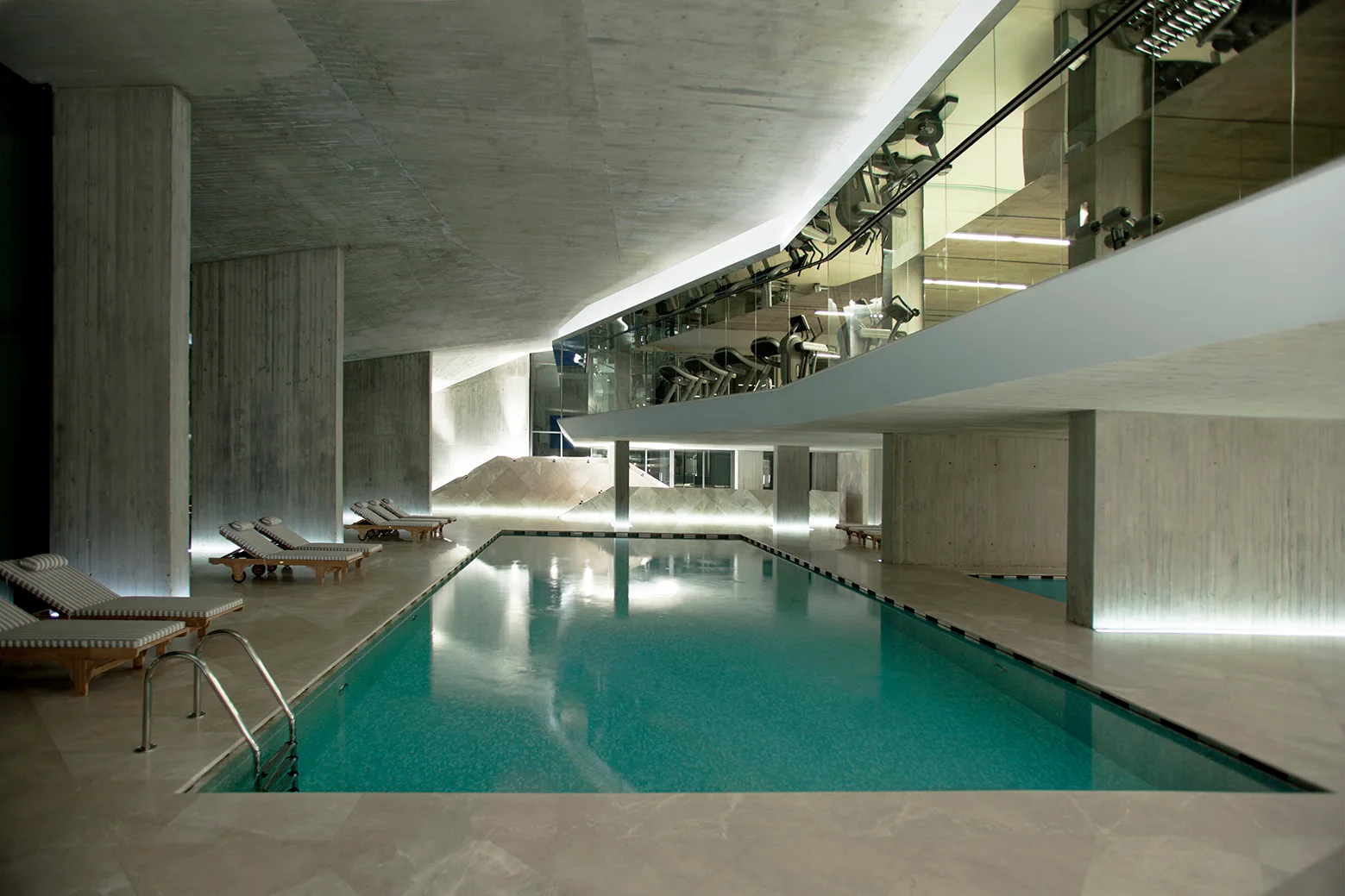 A modern indoor swimming pool surrounded by textured board-formed concrete walls and columns with an elevated gym mezzanine visible behind a glass railing.