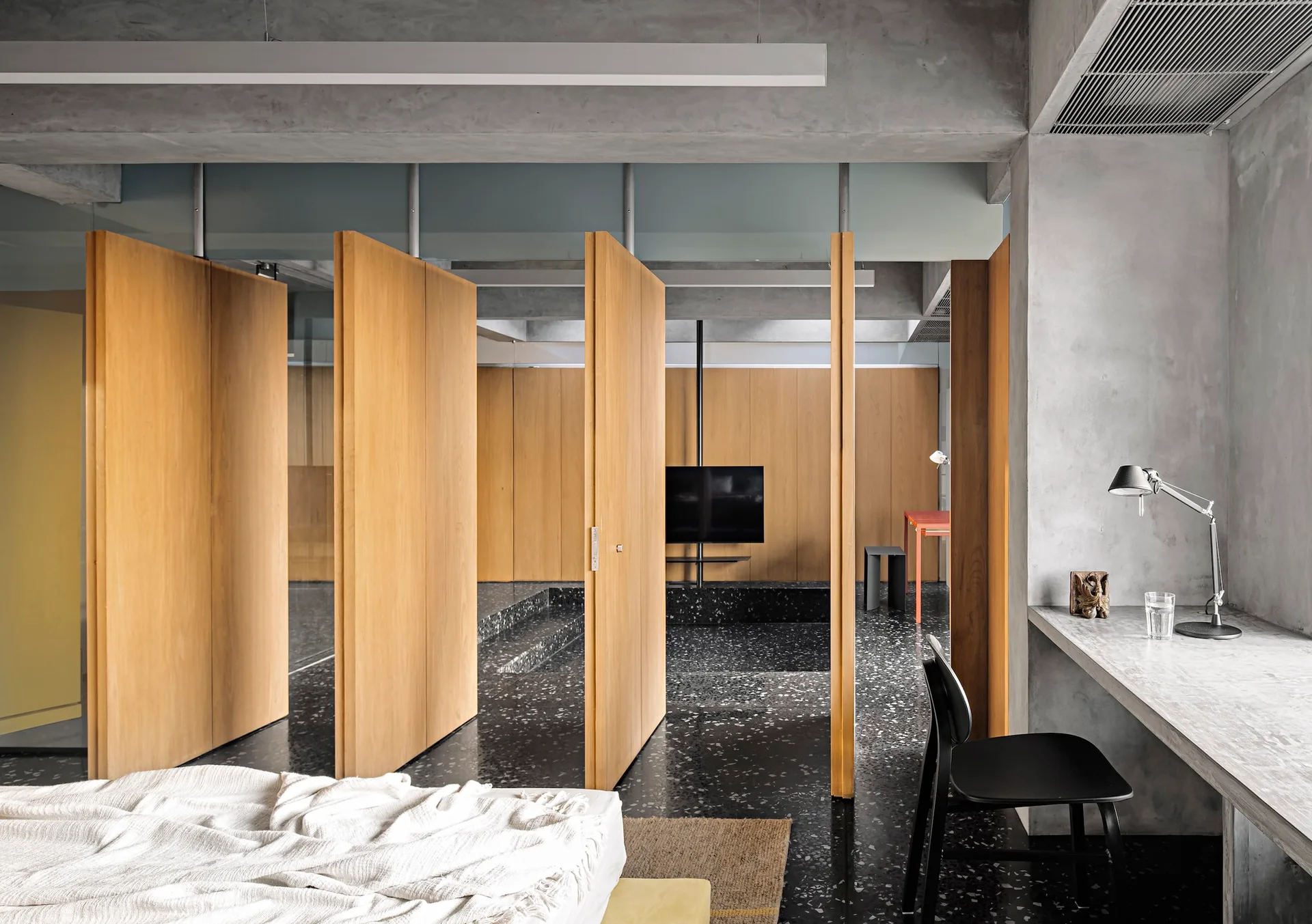 A modern bedroom features tall rotating wood panels separating the sleeping area from a back room with speckled terrazzo floors and a concrete desk.